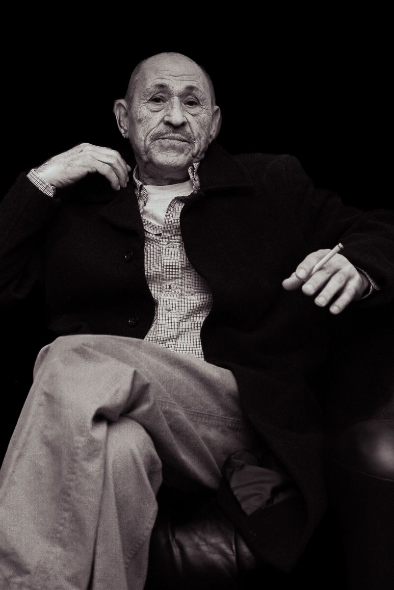 Black and white portrait of an elderly person seated in a relaxed pose wearing a checkered shirt and dark jacket.