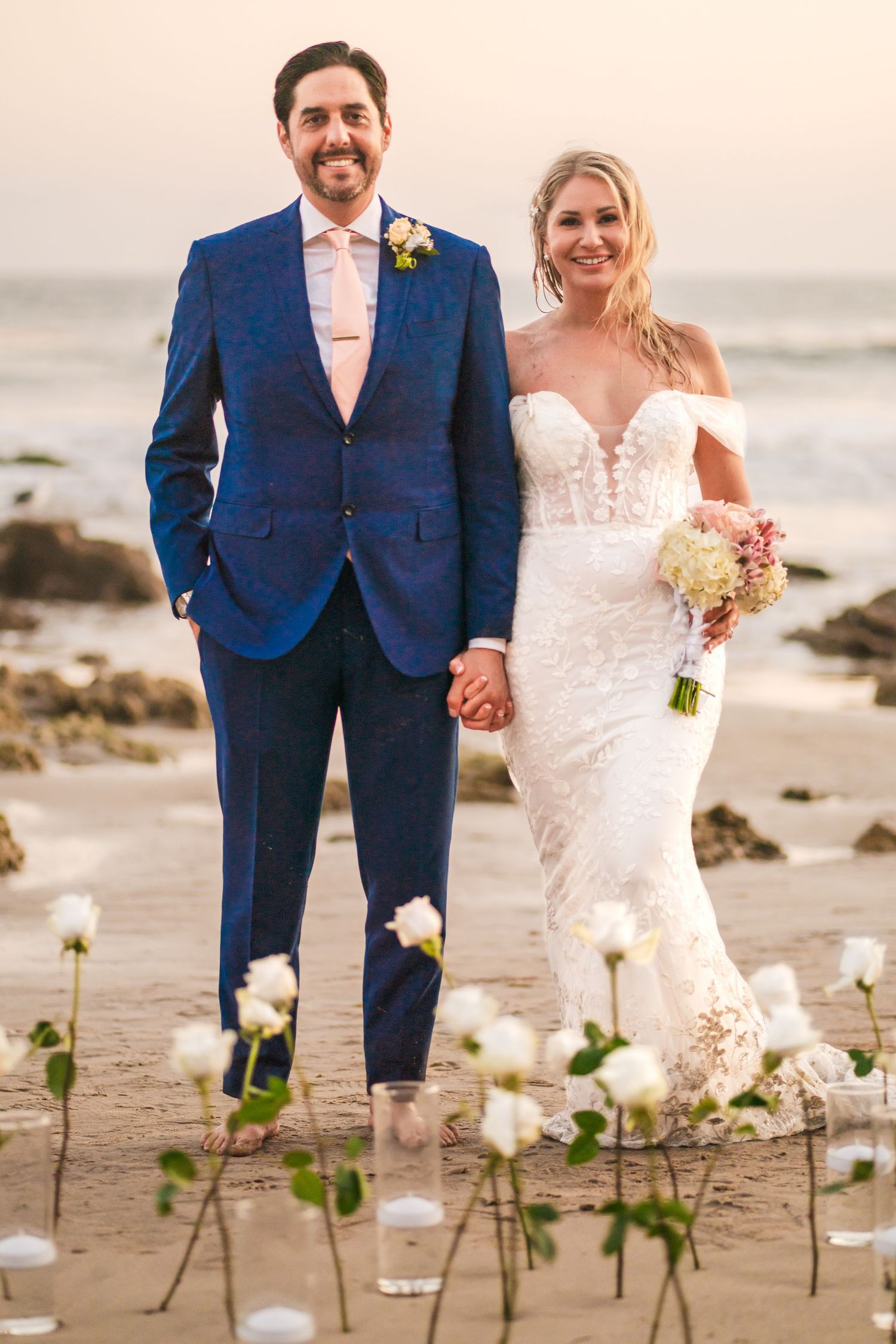 A couple in wedding attire poses together on a beach with white roses scattered in the sand at sunset.