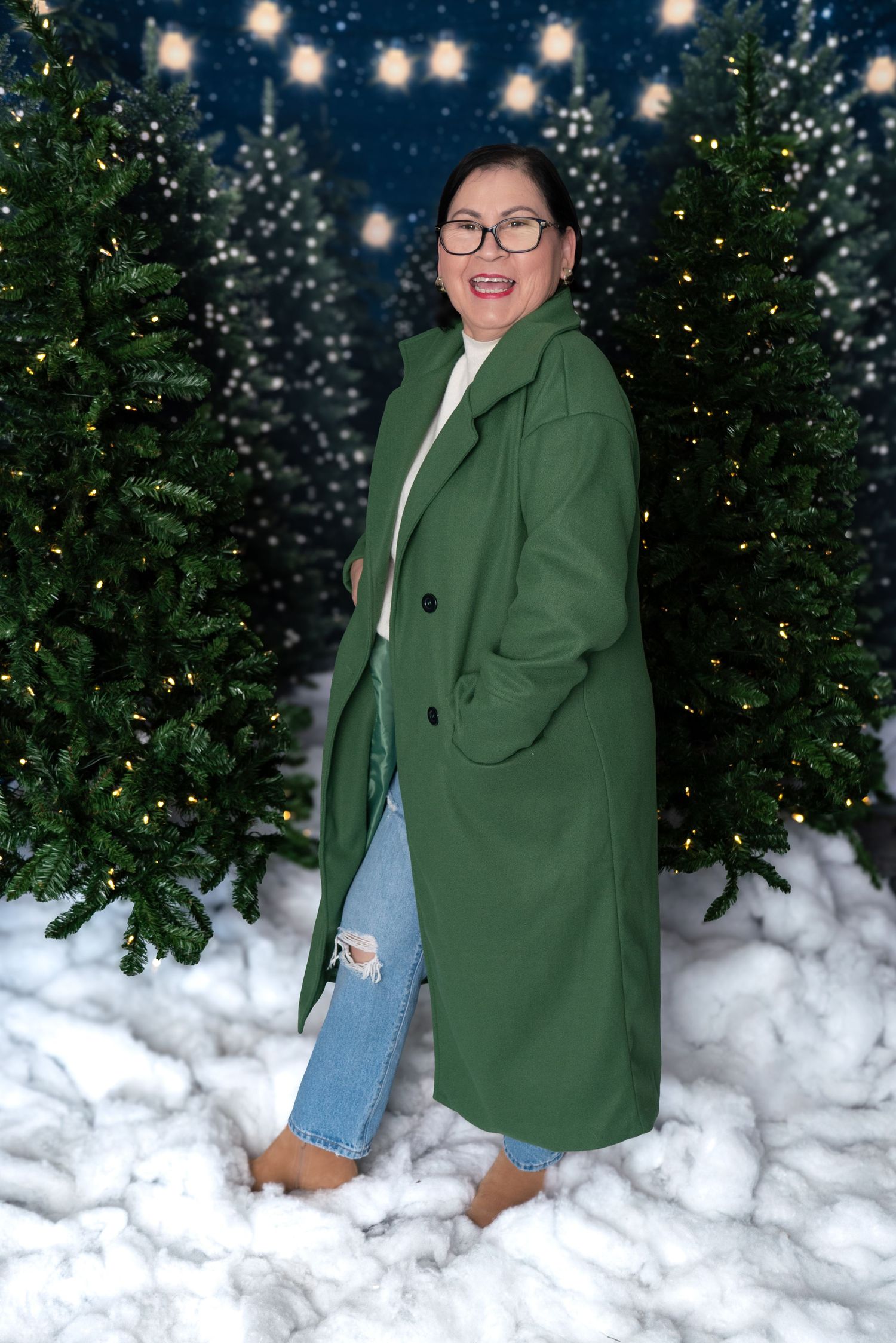 Person wearing a green coat and jeans poses playfully in the snow with Christmas lights and trees in background.