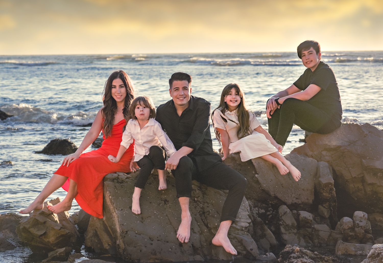 Family relaxing on coastal rocks during golden hour with ocean waves crashing in background.