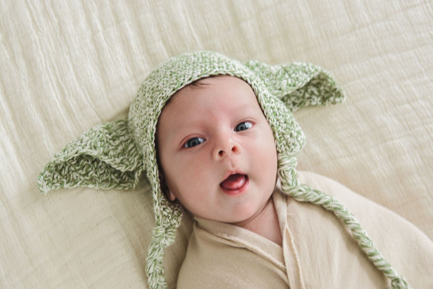 Baby wearing mint green knit bonnet lying on white bedding.