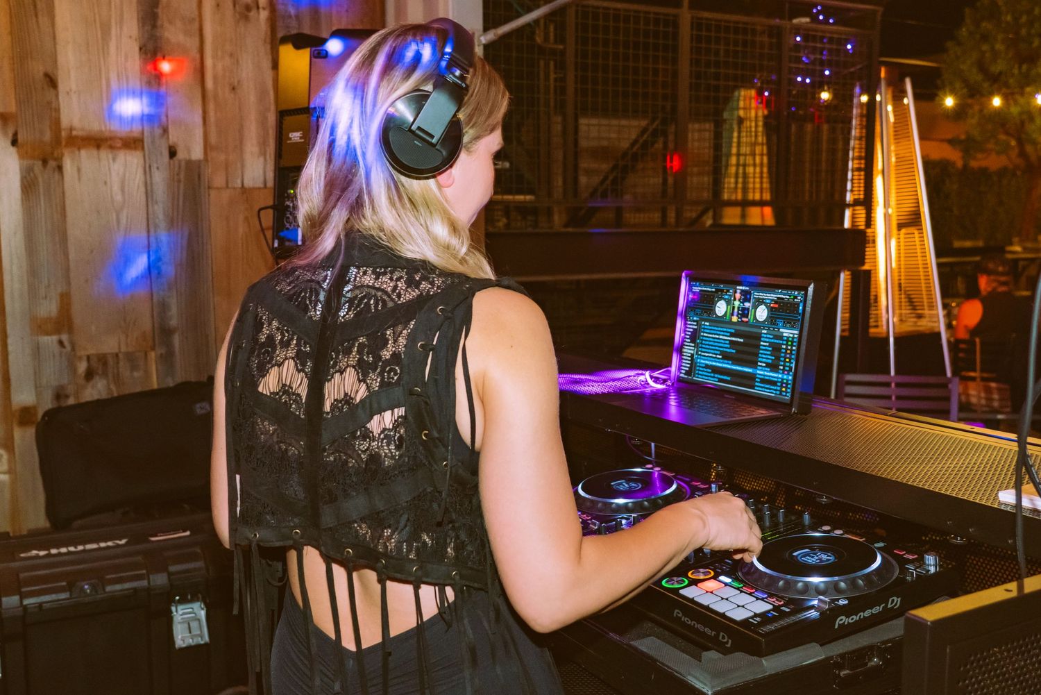 Female DJ wearing headphones mixing music on turntables at an outdoor evening event with purple lighting.