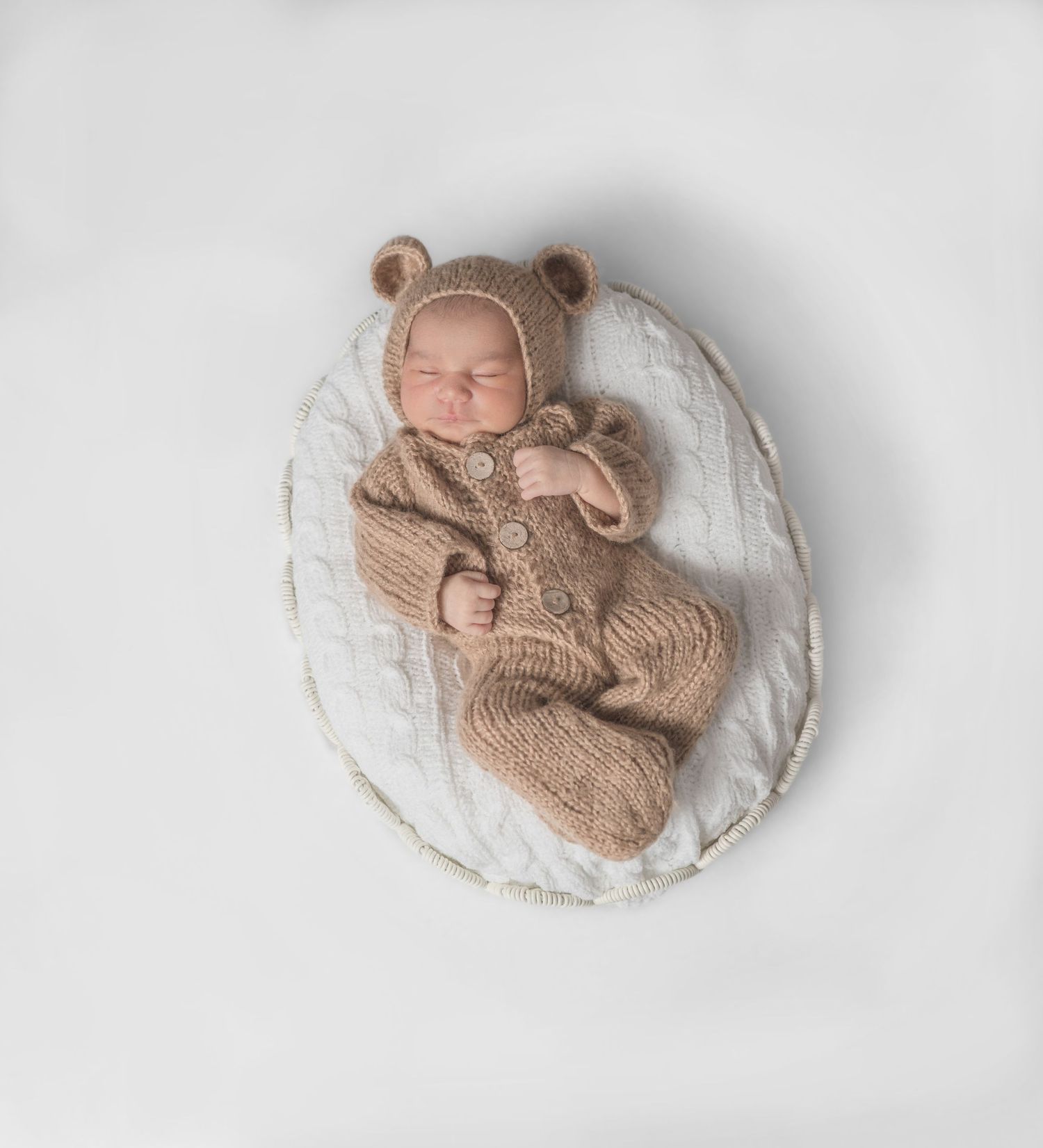 A sleeping newborn baby wrapped in soft beige knit fabric lying on a white textured backdrop.