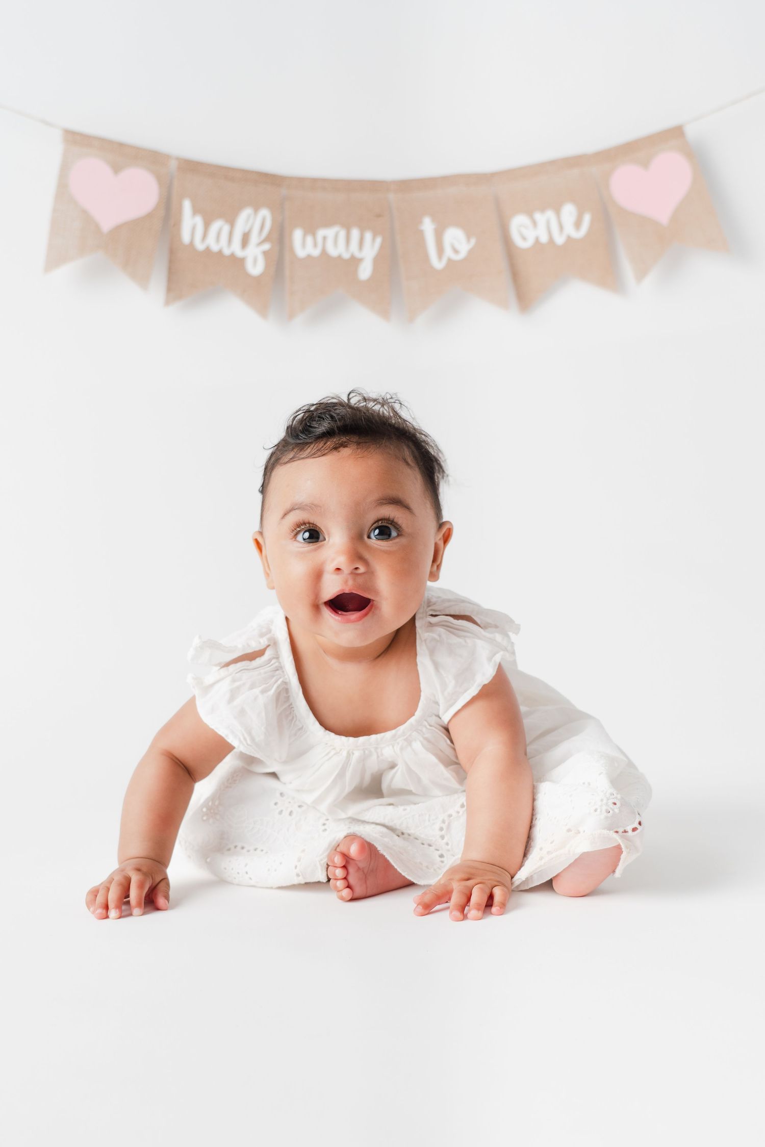Baby in white dress crawls under 'half way to one' burlap banner with pink hearts.