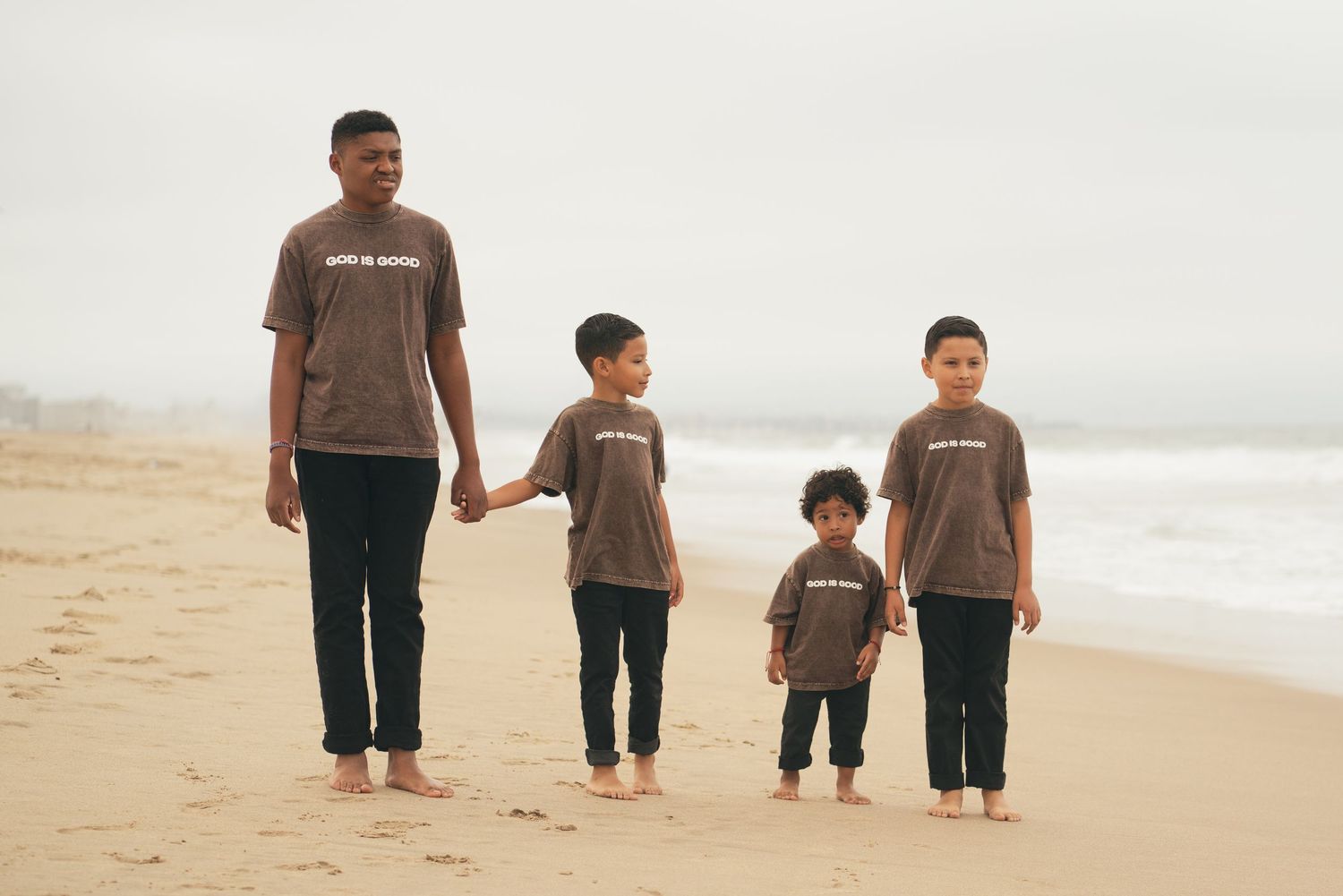 A man and three young boys wearing matching brown t-shirts walk hand in hand on a sandy beach.