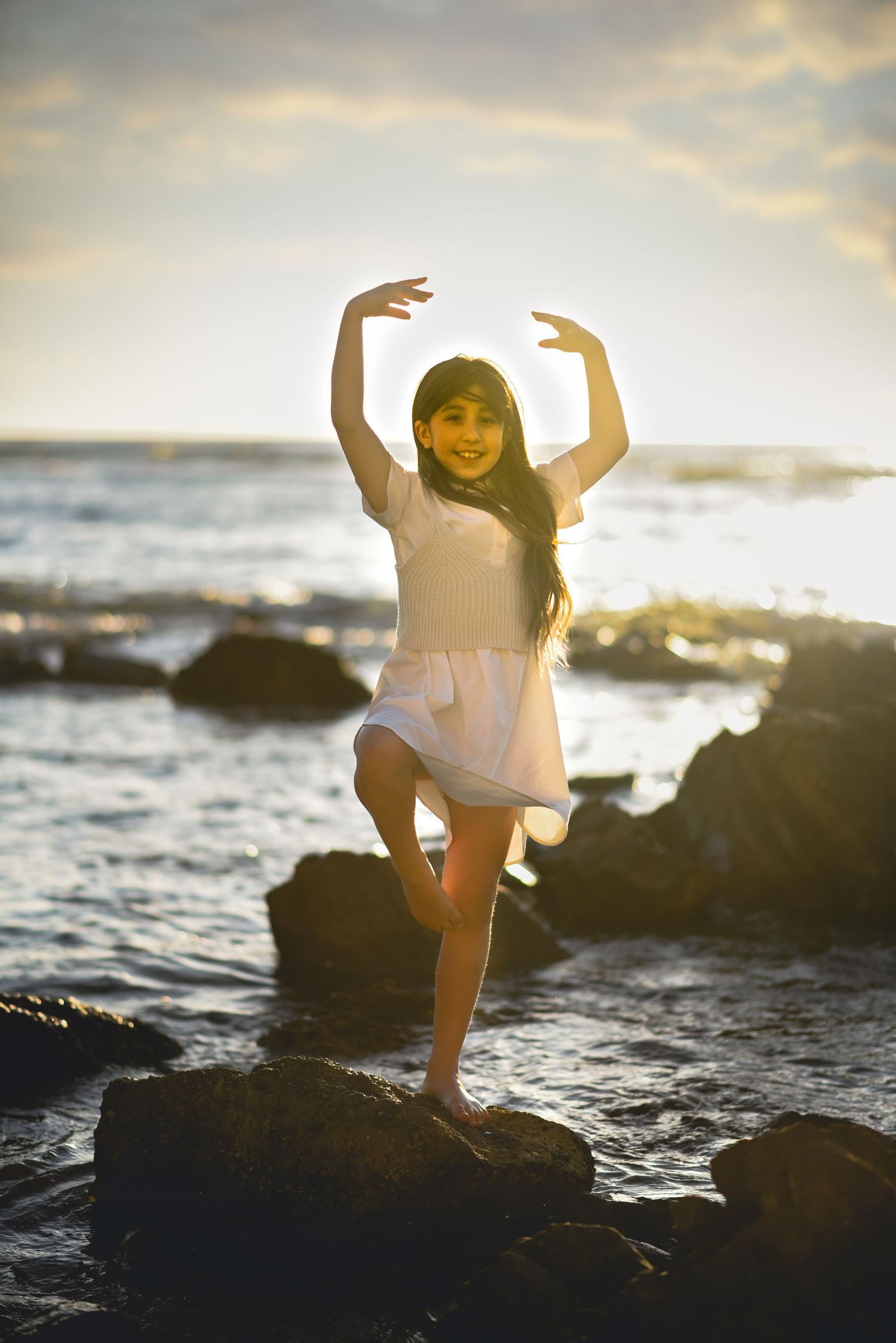 A young dancer poses gracefully on rocks by the ocean at sunset.