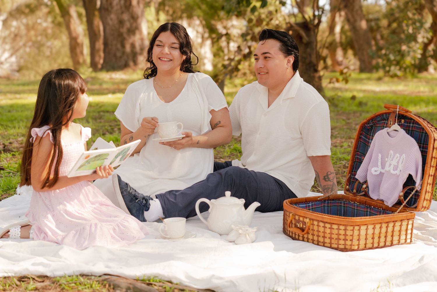 A family enjoys a picnic together on a white blanket in a sun-dappled outdoor setting.