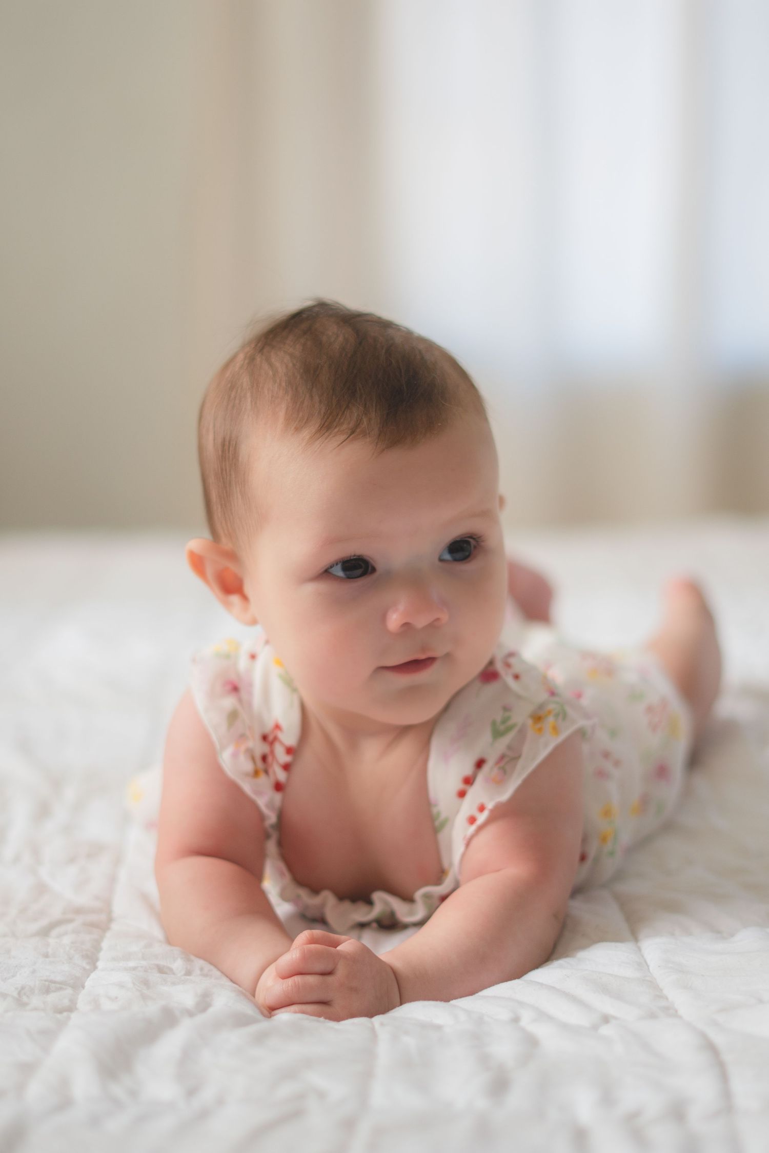 A cute baby in a floral romper doing tummy time on a white quilted blanket, looking up curiously.