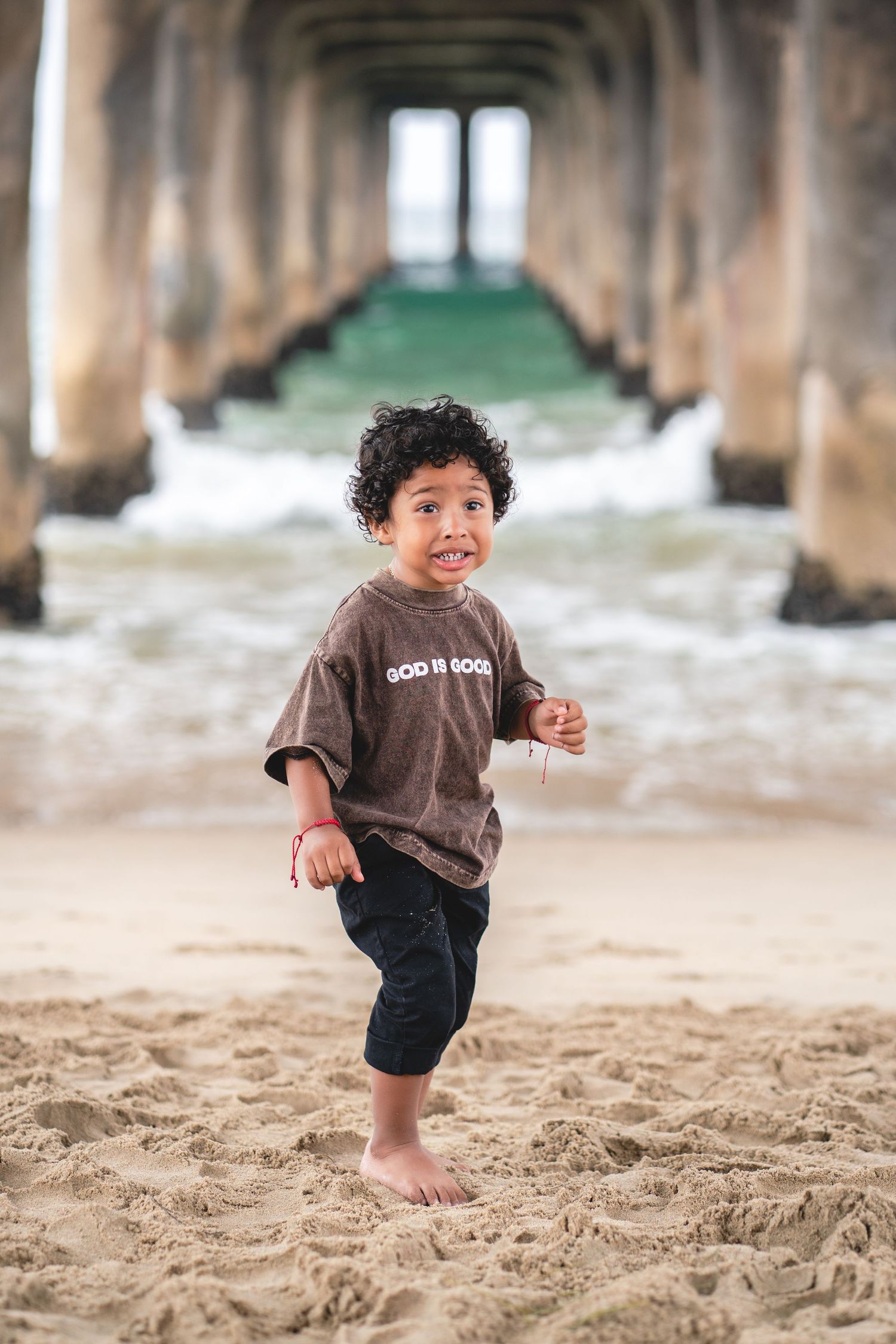 Happy young boy in brown God Is Good shirt running barefoot on sandy beach under a pier.