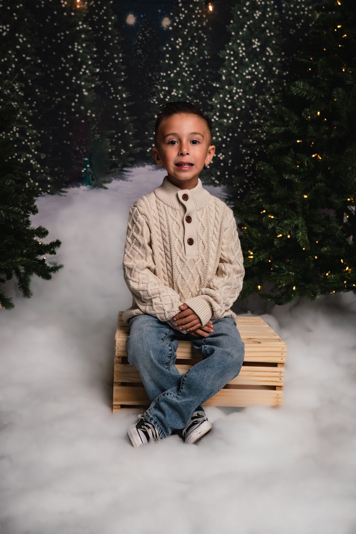 Portrait photo with festive Christmas backdrop featuring cable knit sweater and jeans against sparkly pine trees and fog.