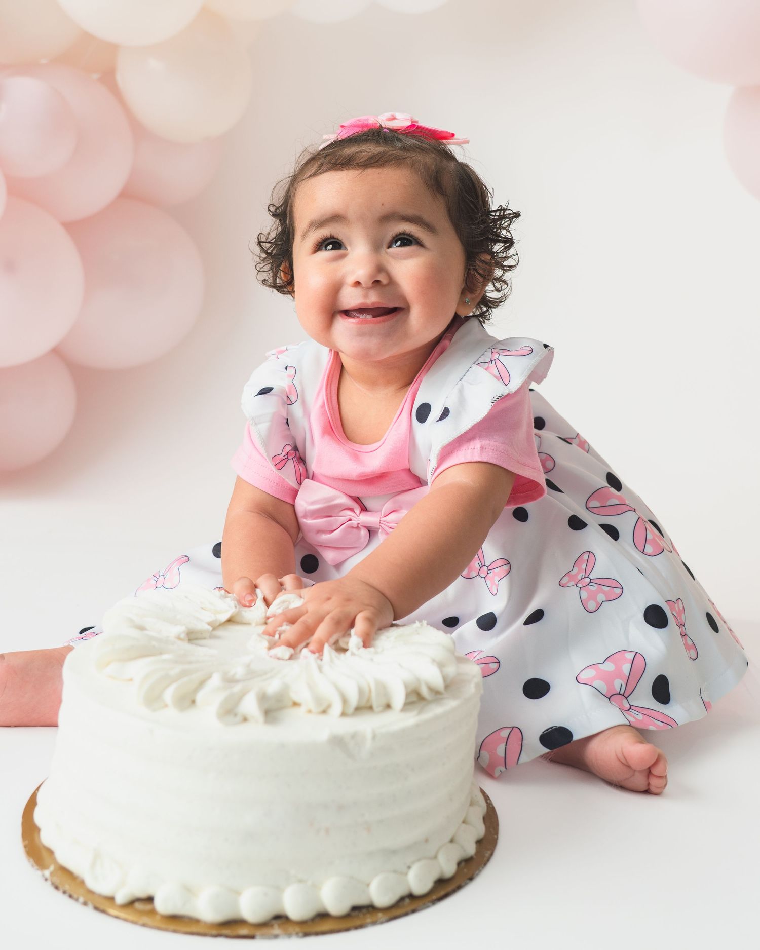 Baby in polka dot dress reaches for white frosted cake during cake smash photo session.
