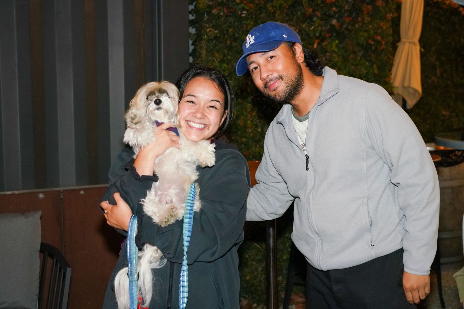 Smiling couple posing outdoors at night holding an adorable small fluffy dog on a leash.