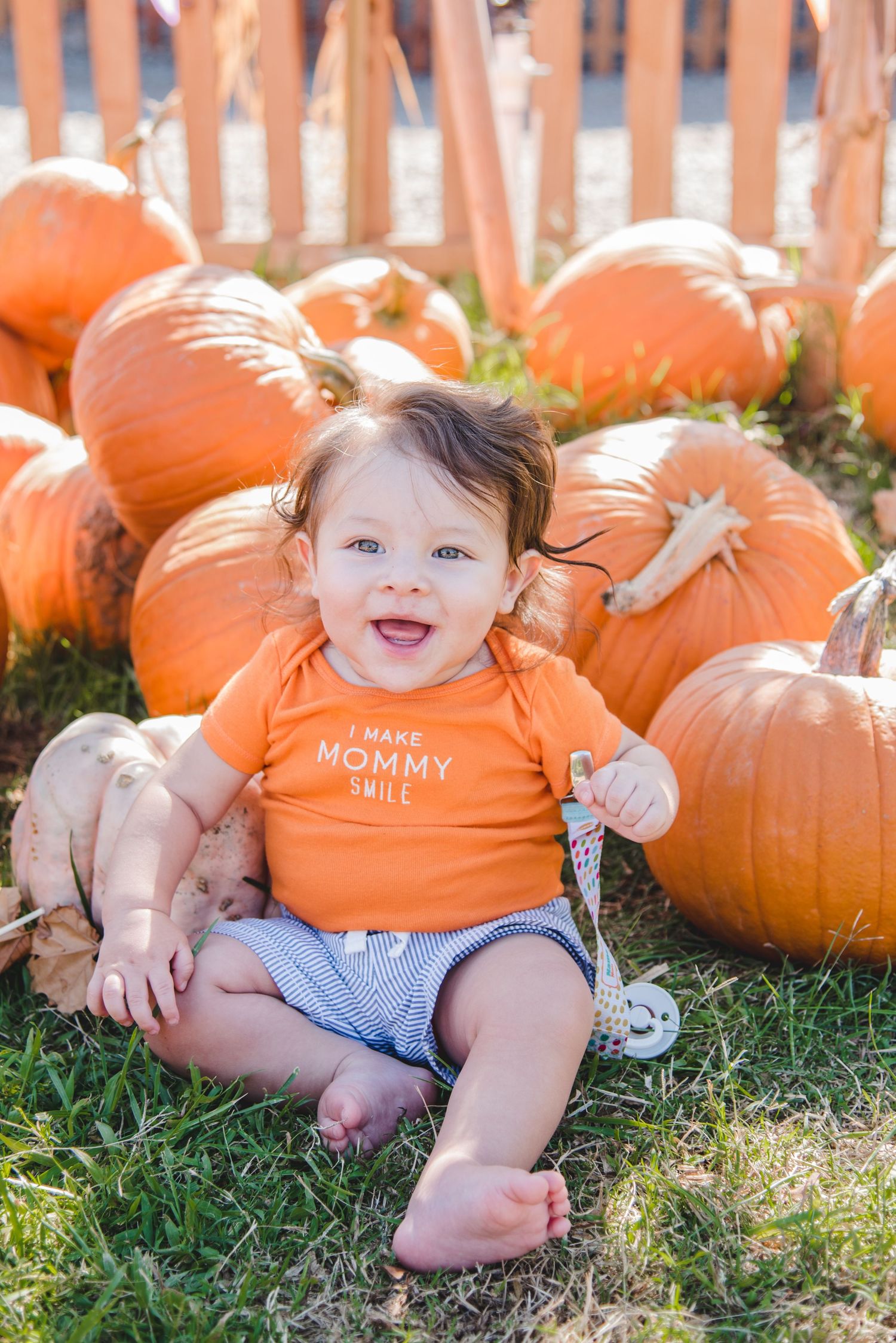 Baby wearing orange shirt sits among large orange pumpkins in grass at pumpkin patch during fall season.
