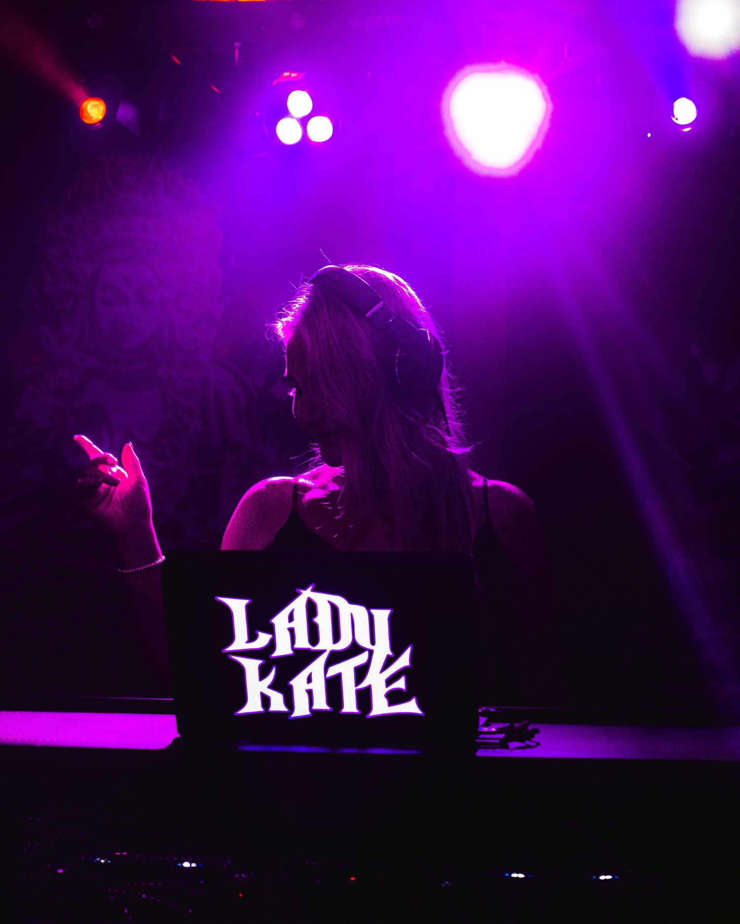 DJ Lady Kate performing on stage under purple and pink stage lights with headphones and illuminated DJ booth.