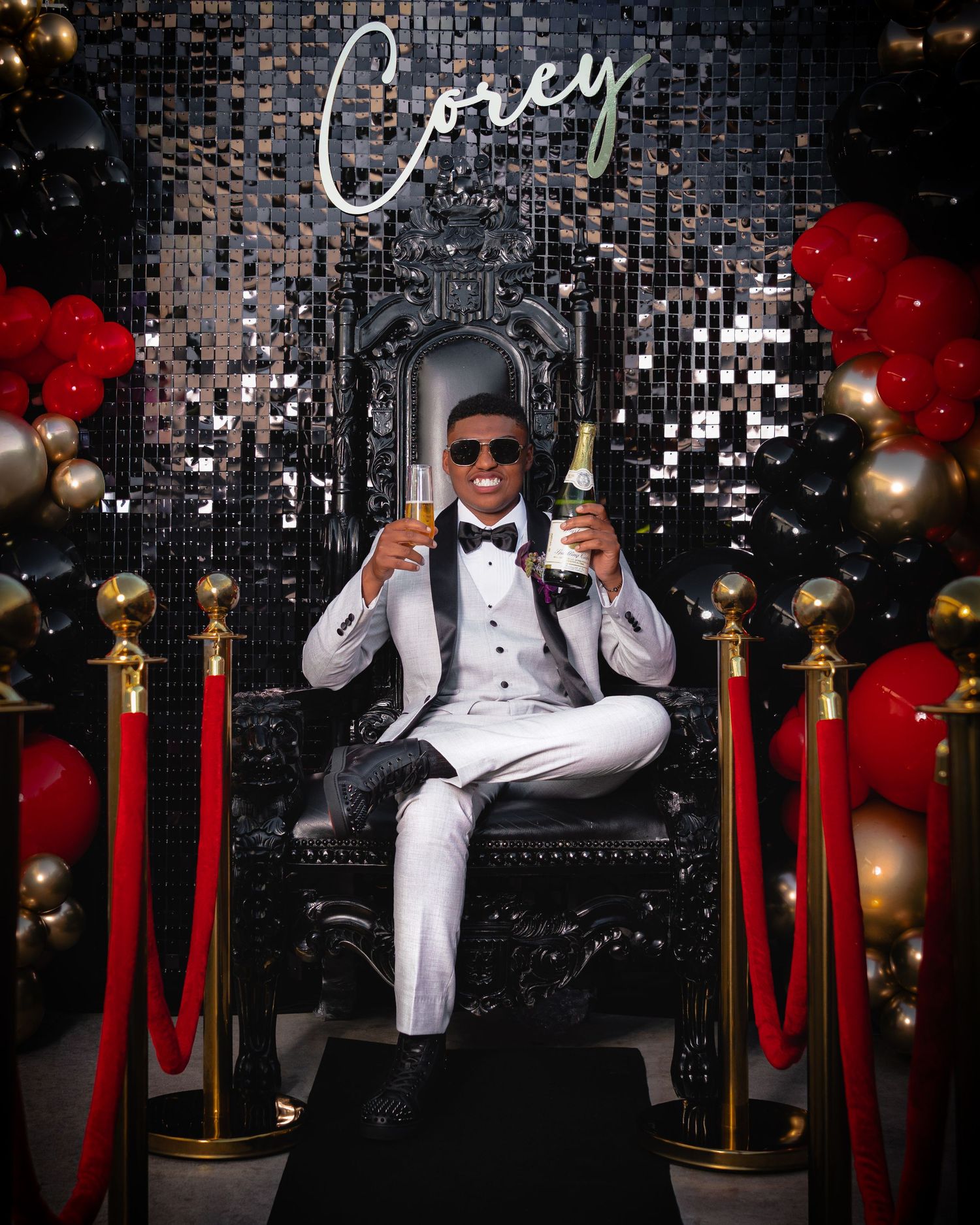 Man in white tuxedo sitting on ornate throne chair holding champagne at elegant Corey celebration event.
