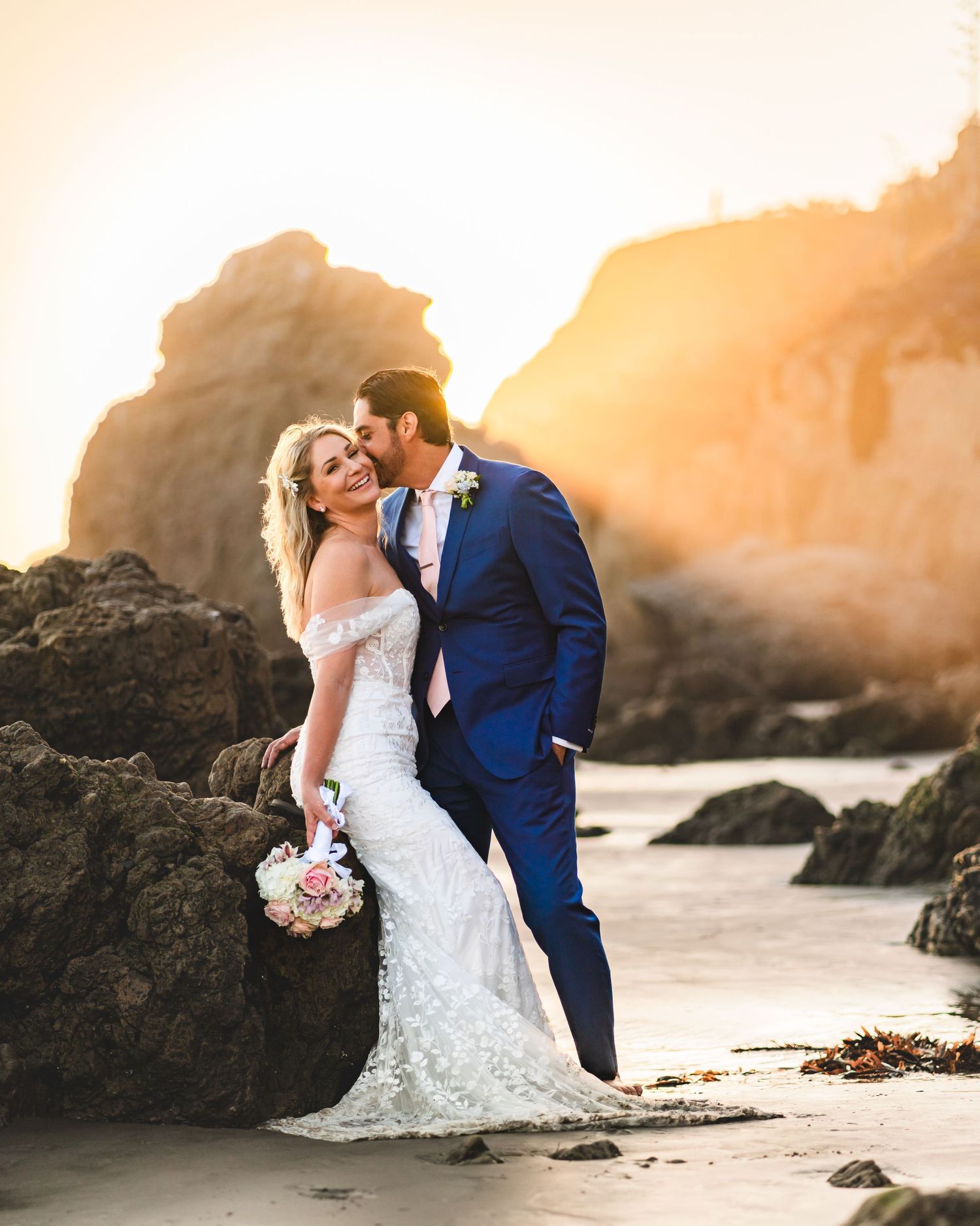 Couple in wedding attire embrace on a beach at sunset with golden light illuminating rocky coastal cliffs.