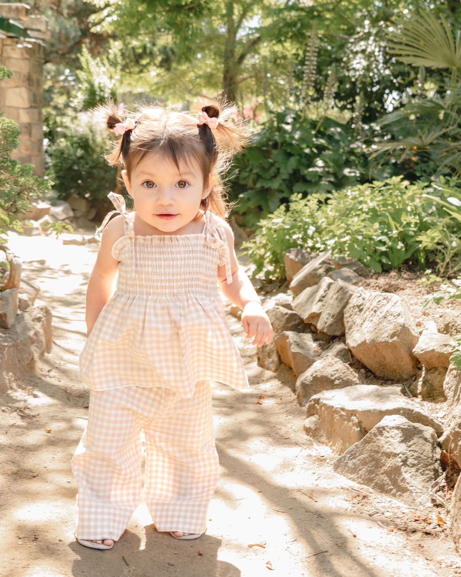 A small child in a peach gingham dress plays outdoors in dappled sunlight among rocks and greenery.