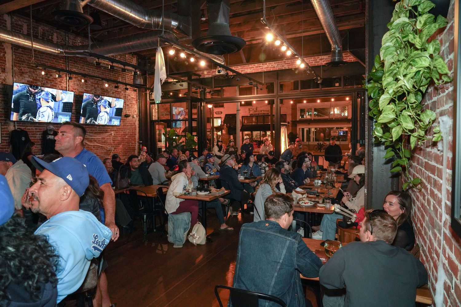 Crowded restaurant bar with exposed brick walls, TV screens, and lush greenery decor during a busy evening gathering.