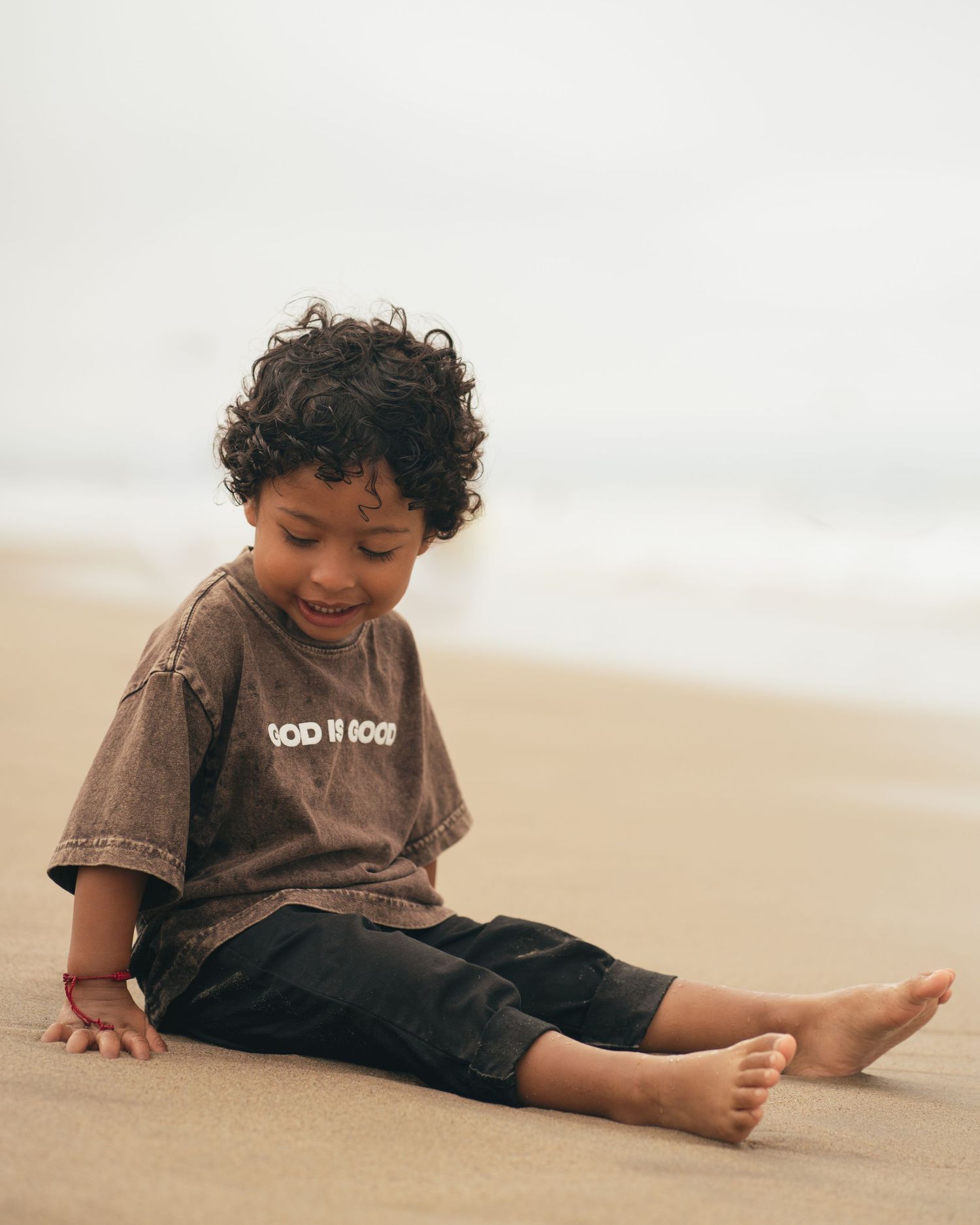 Adorable curly-haired toddler in a brown t-shirt sitting barefoot on a sandy beach, smiling softly.