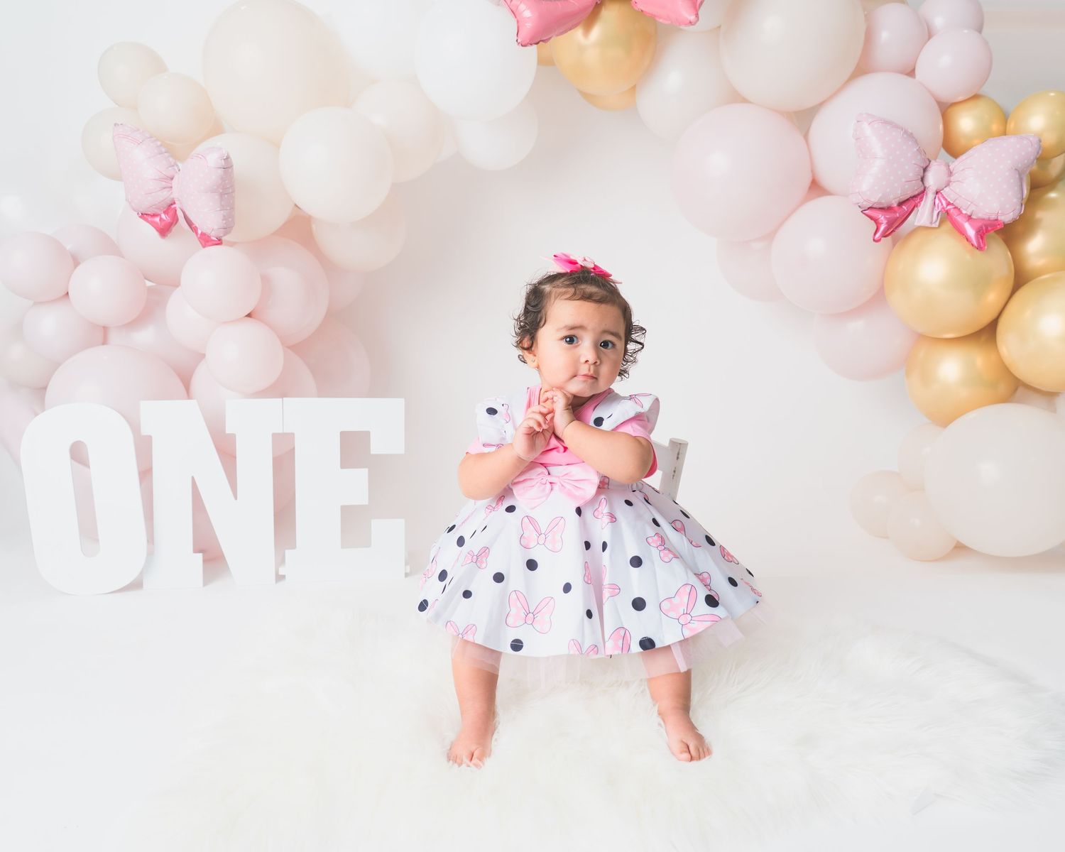 Baby in polka dot dress lies on white floor with pastel balloon decorations.