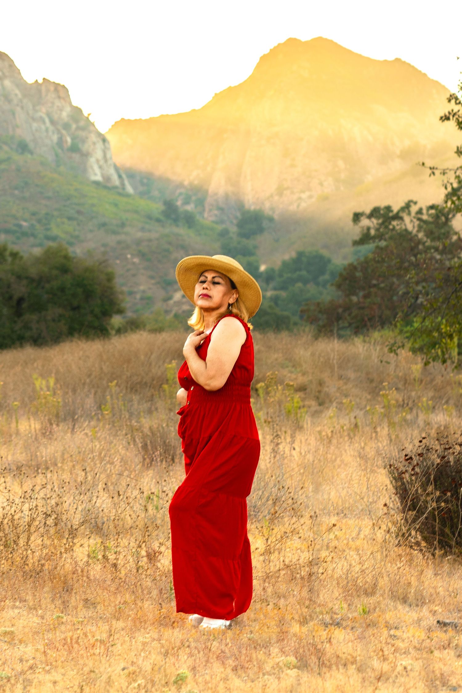 Figure in a flowing red dress and straw hat poses gracefully against golden hills and mountain backdrop during sunset.