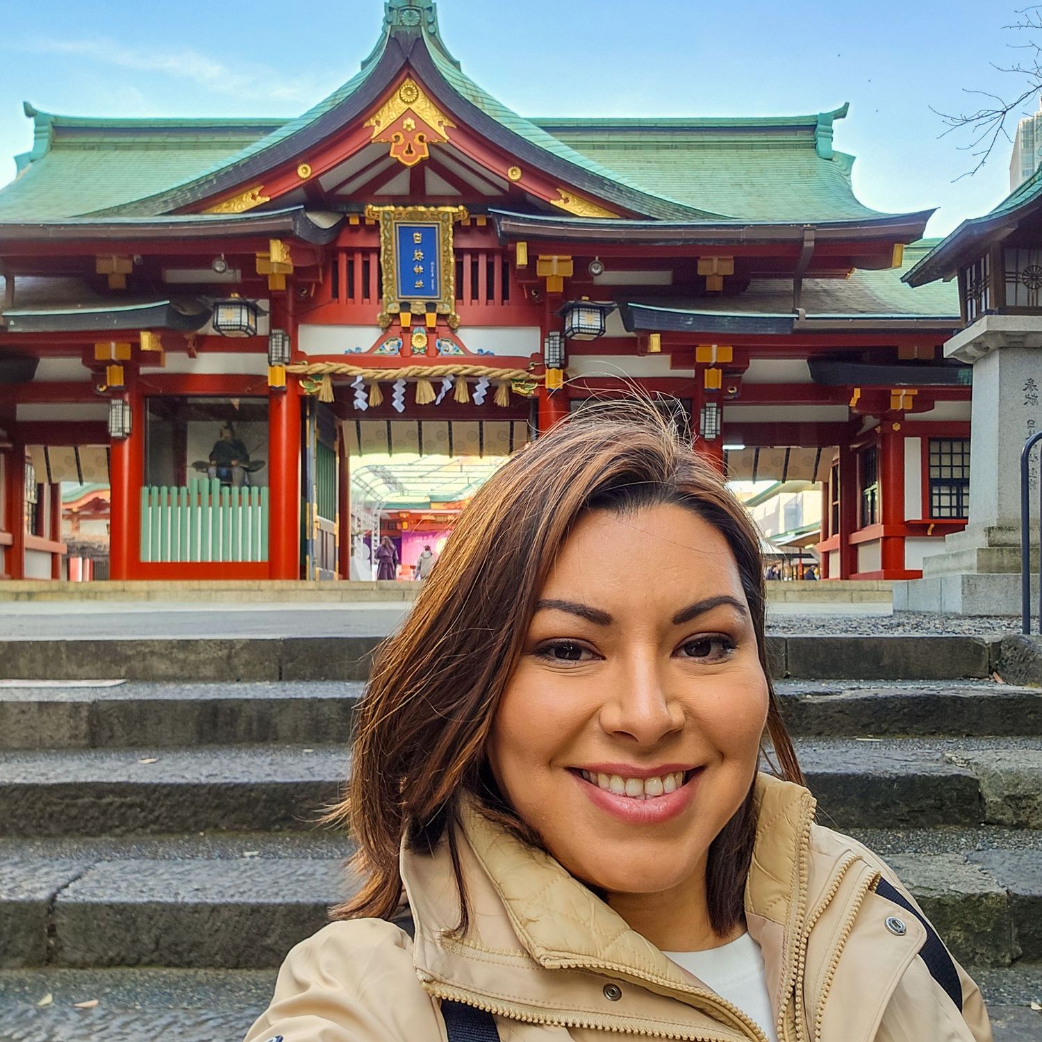 A visitor wearing a beige coat smiles while exploring a traditional Japanese shrine with red and green architecture.