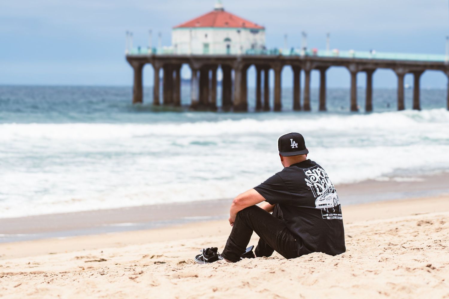 A person lies on their stomach on a sandy beach with ocean waves and a wooden pier in the background.