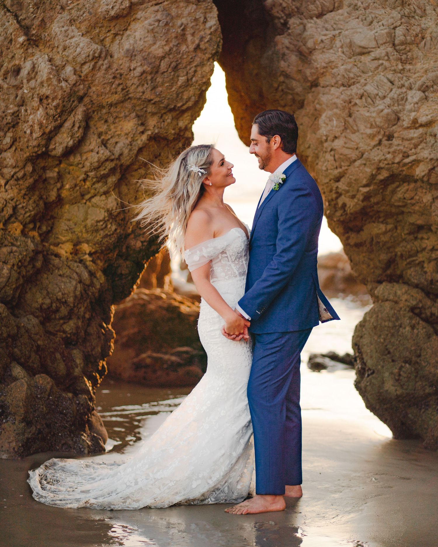 A romantic wedding photo at sunset showing a couple embracing under a natural rock arch formation on a beach.