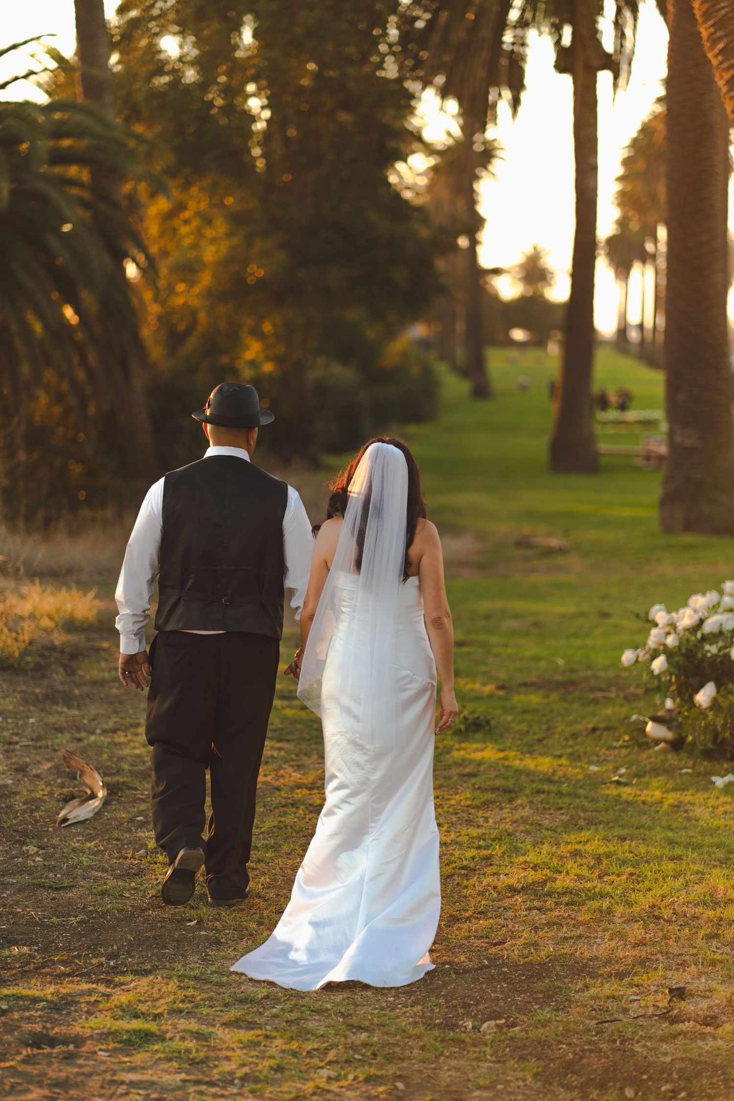 Bride and groom walking hand in hand through a sunlit tree-lined path, bride in white gown with veil, groom in black vest.