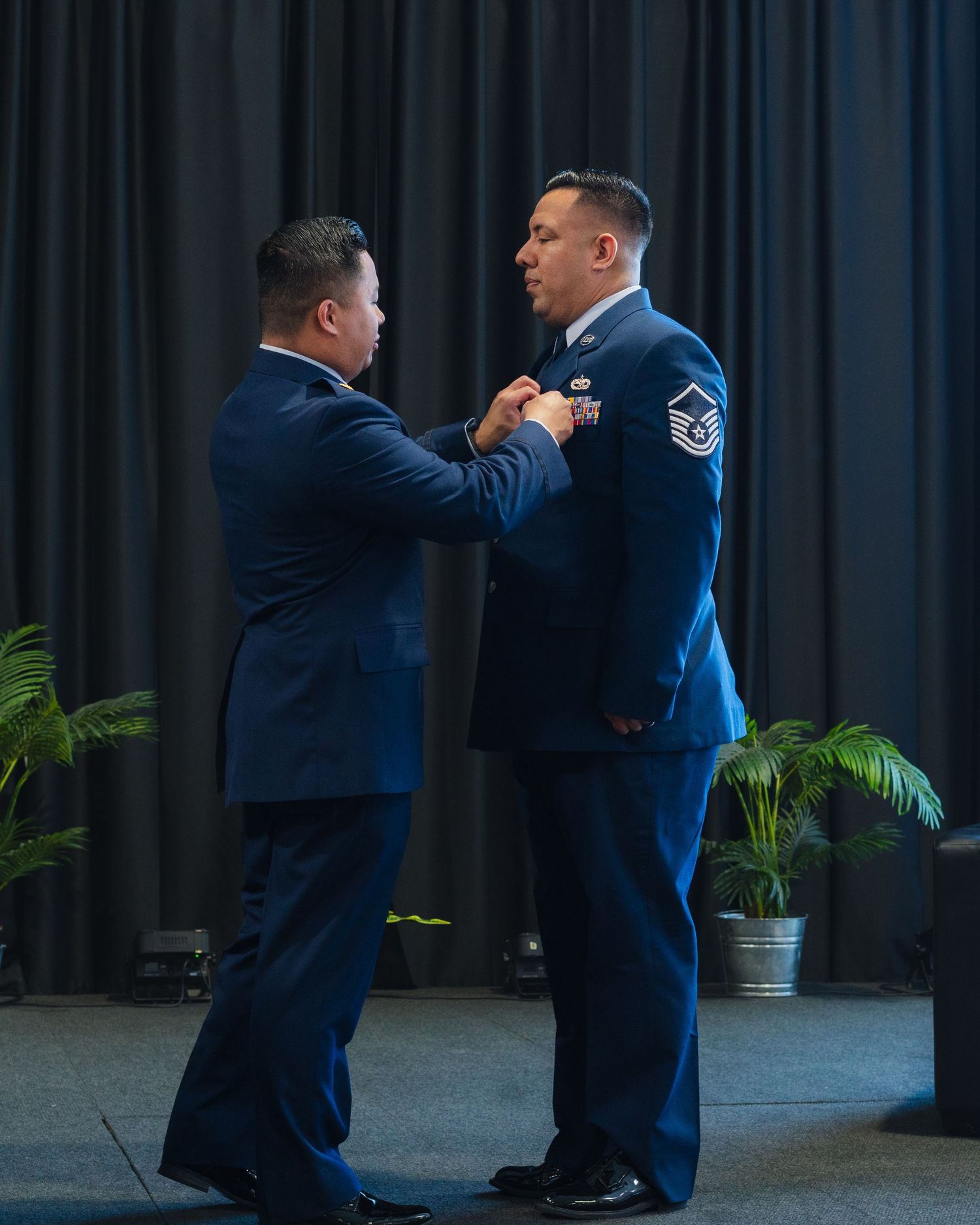 Military ceremony with two airmen in dress blues, one pinning a medal on the other during a formal promotion event.