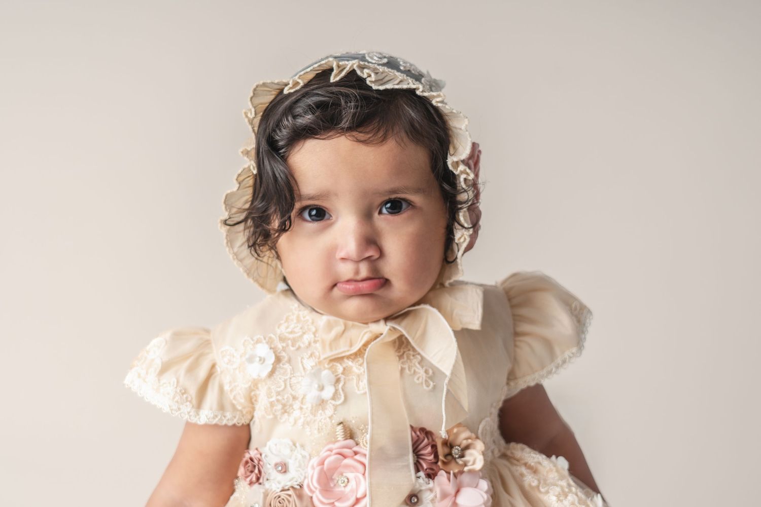 A toddler wearing a delicate cream-colored lace dress and headband against a neutral backdrop.