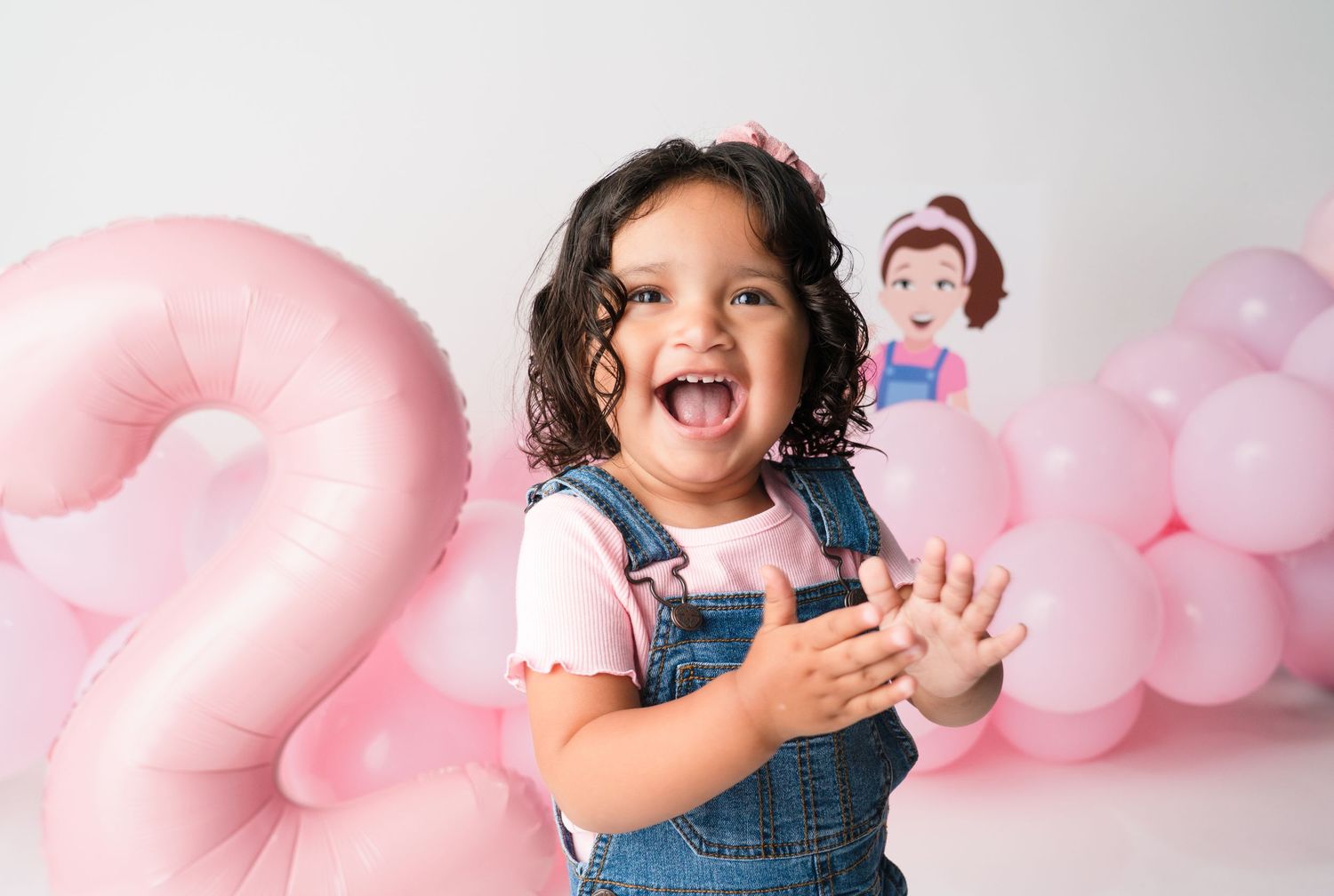 A child in denim overalls plays with pink balloons against a white backdrop with playful expressions.