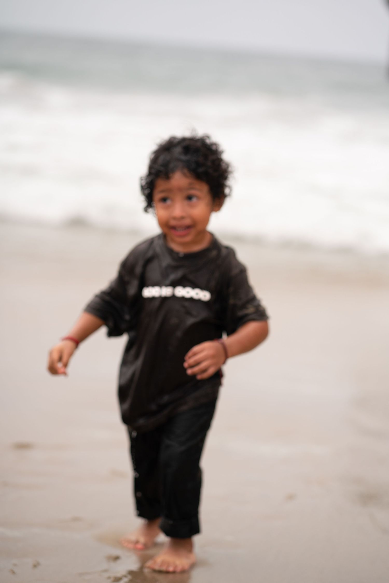 Smiling toddler in black God Is Good shirt walking barefoot on a sandy beach near the ocean.