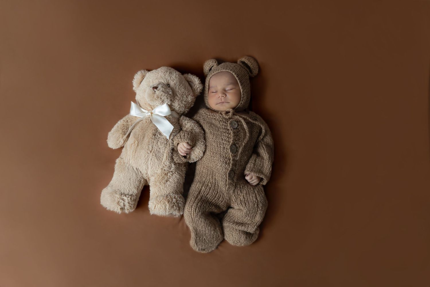 A newborn in a brown knitted bear outfit lies next to a matching teddy bear on brown background.