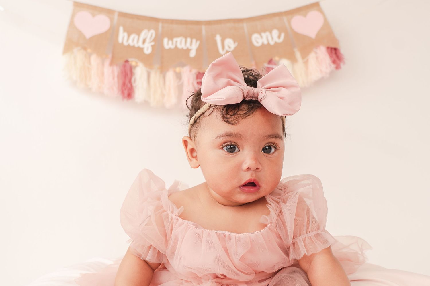 Baby in pink dress with bow under 'half way to one' burlap banner with hearts.
