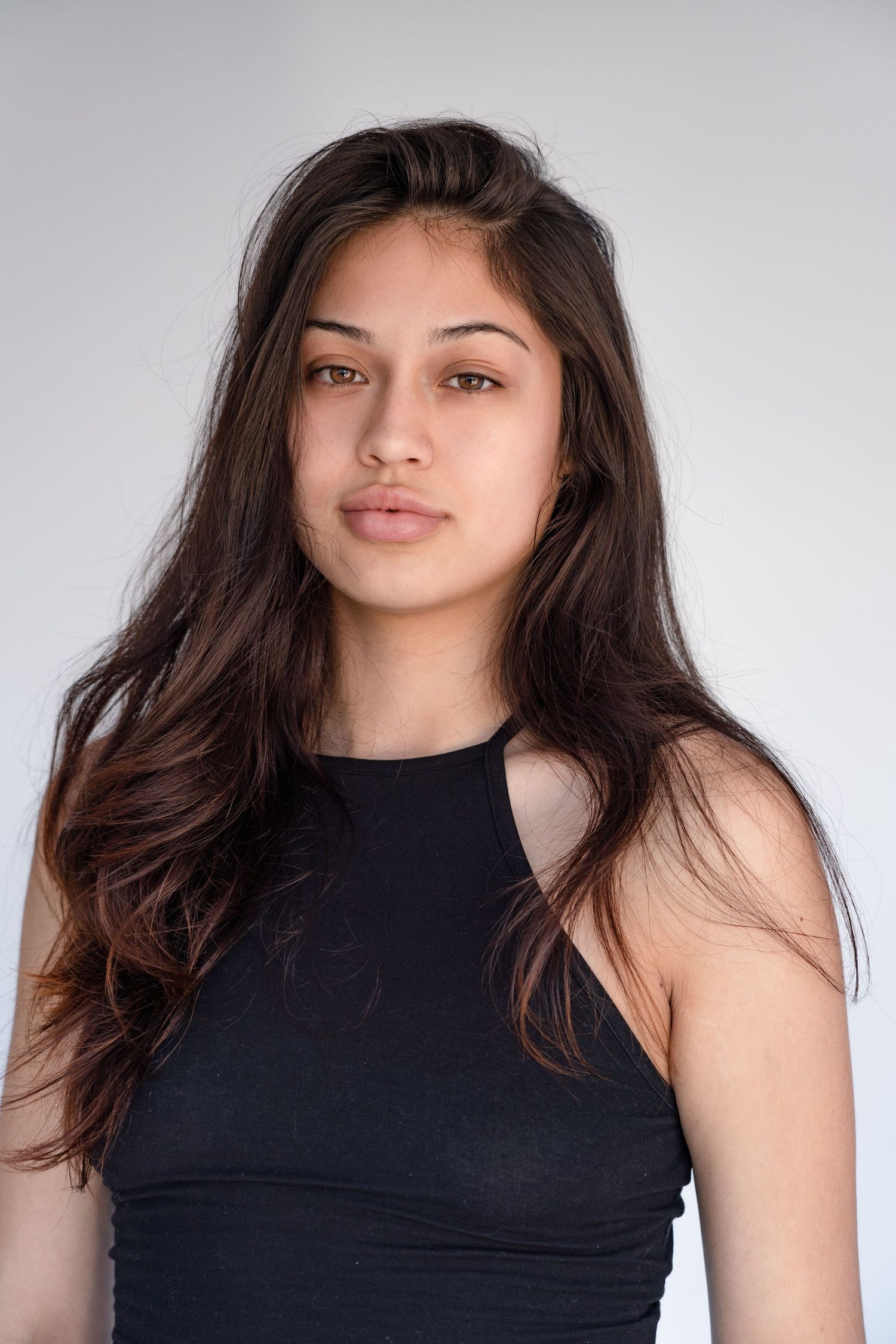 A studio portrait featuring long dark hair and natural makeup against a light background.