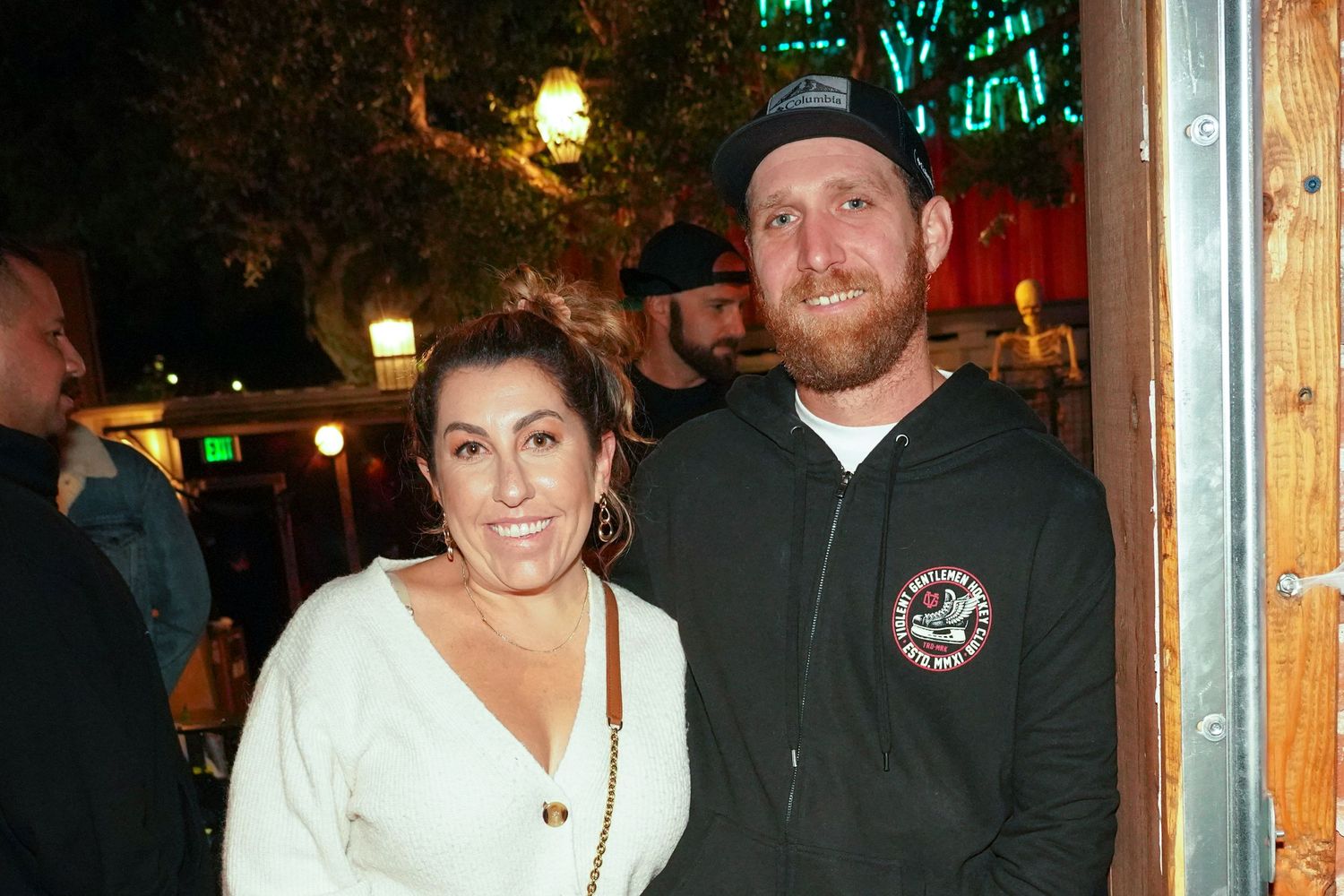 A smiling couple poses outdoors at night, woman in white sweater and man in dark hoodie with cap.
