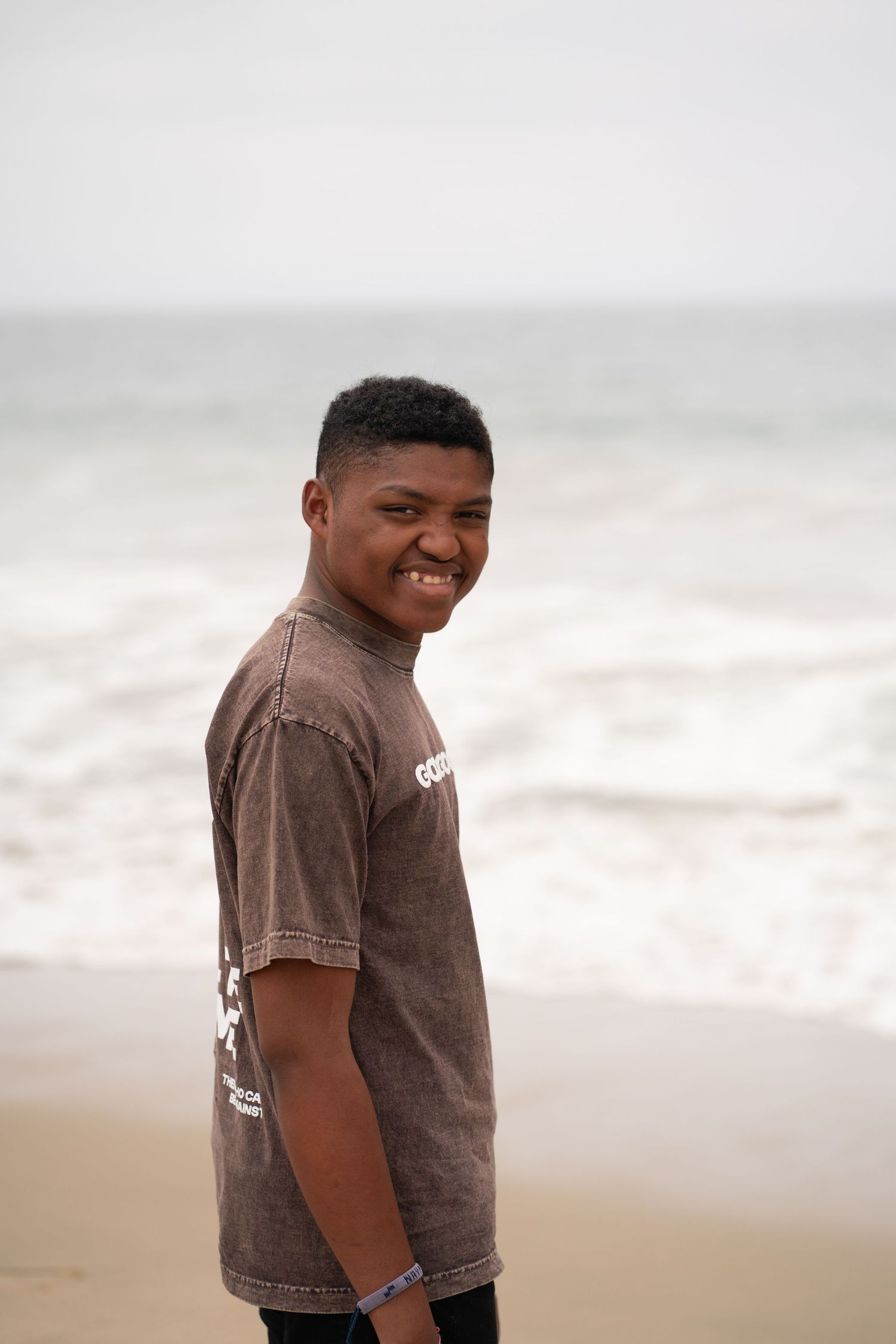 Smiling young man in brown t-shirt standing on a sandy beach with ocean waves in the background.