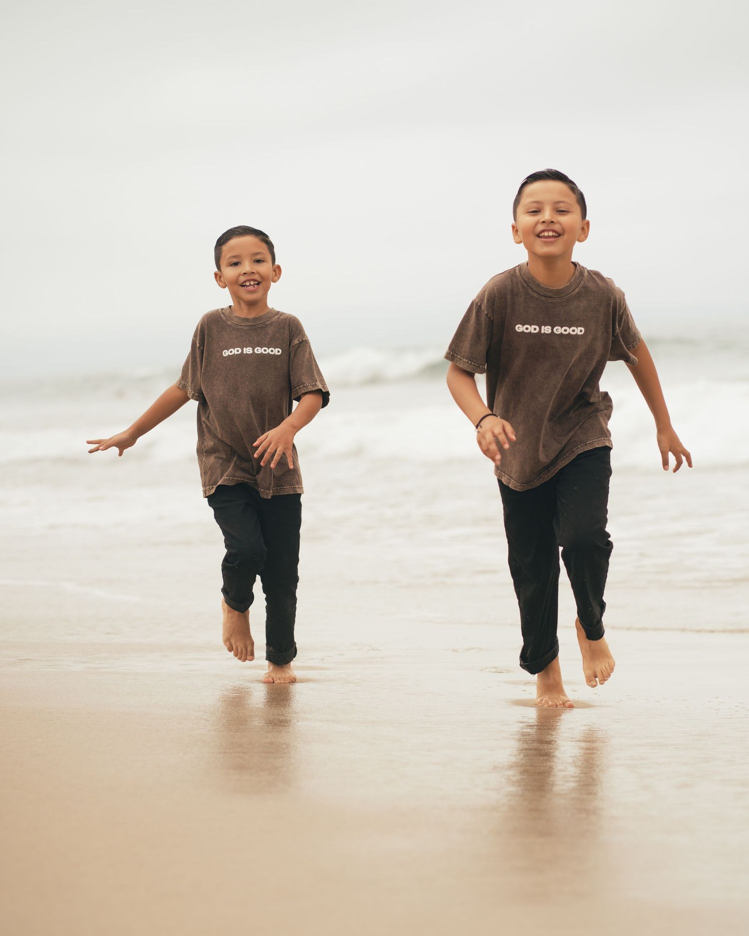 Two young boys in matching brown God Is Good t-shirts run barefoot along a sandy beach smiling.