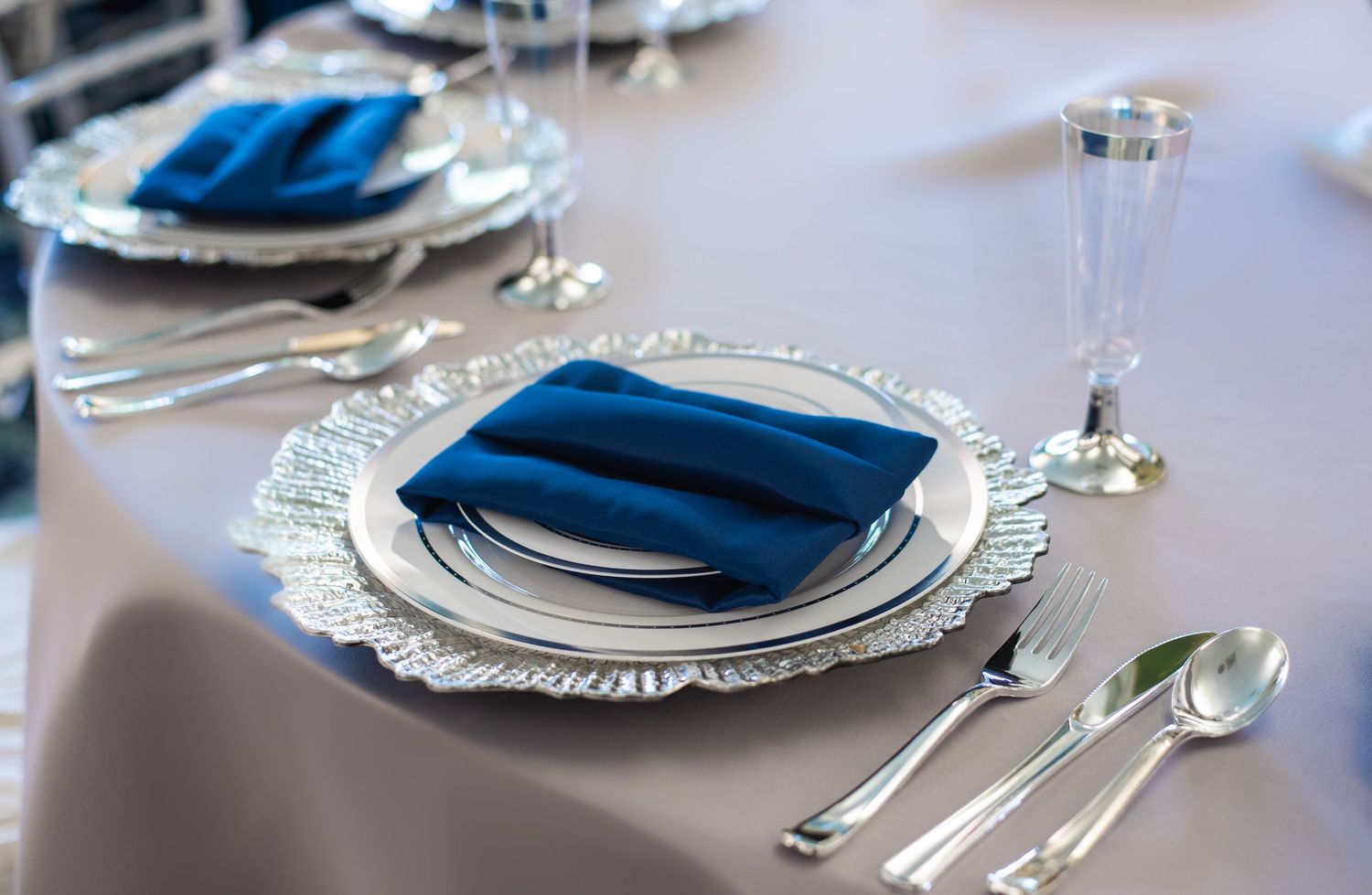 Formal place settings with silver charger plates and navy blue napkins on white tablecloth.