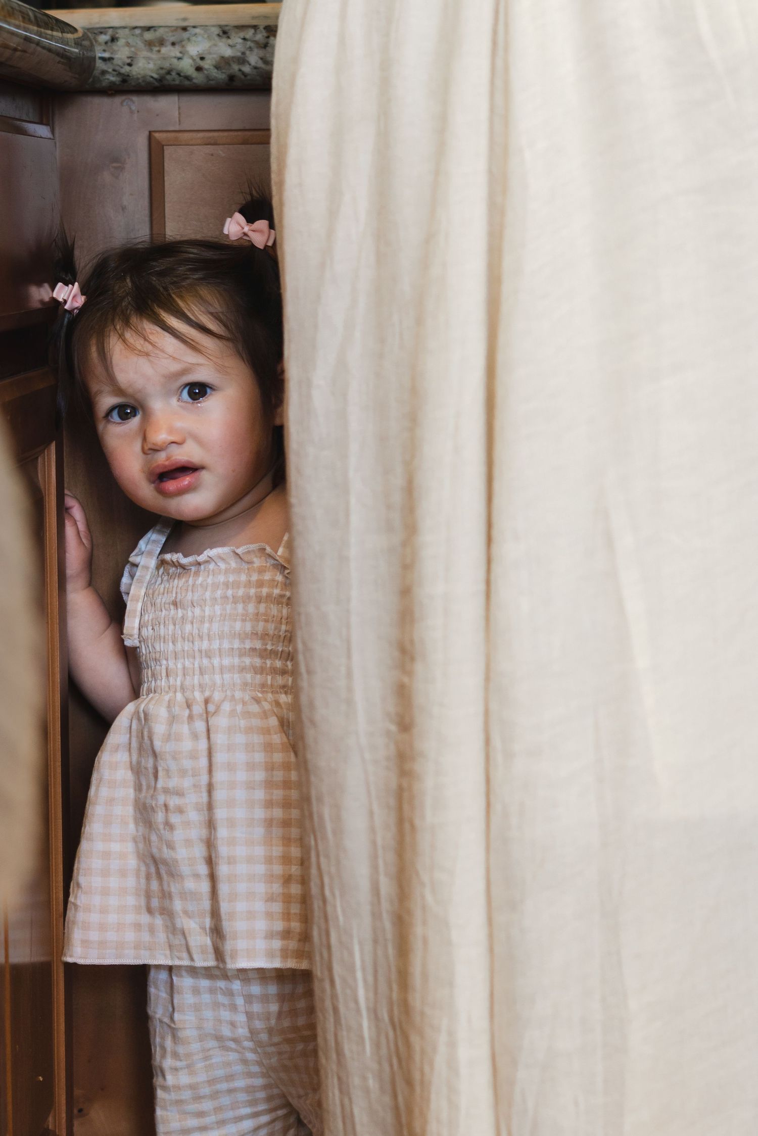 Small child in checkered dress plays peek-a-boo behind white curtains.