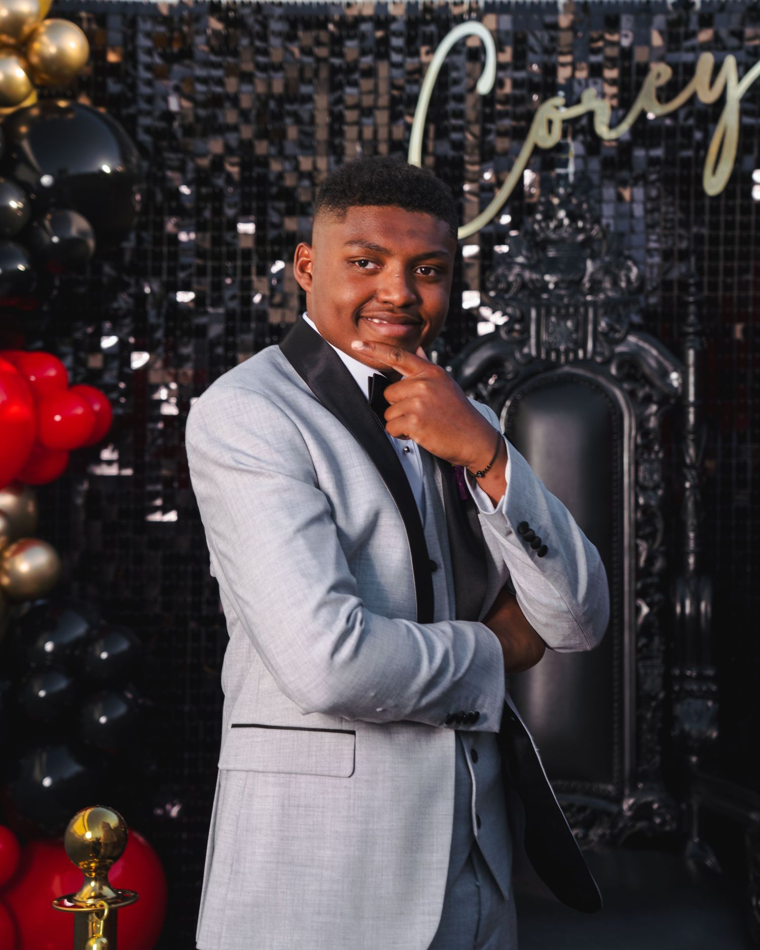 Young man in light gray tuxedo poses confidently at elegant black and gold party backdrop with balloons.