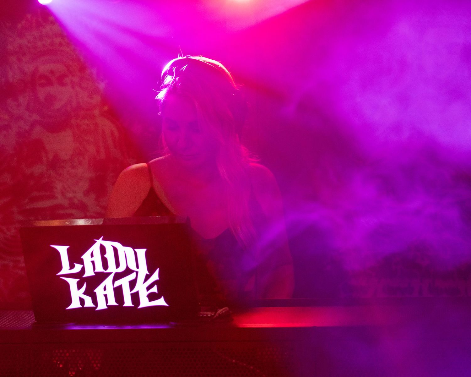 DJ Lady Kate performing under vibrant pink and purple stage lights in a smoky nightclub.