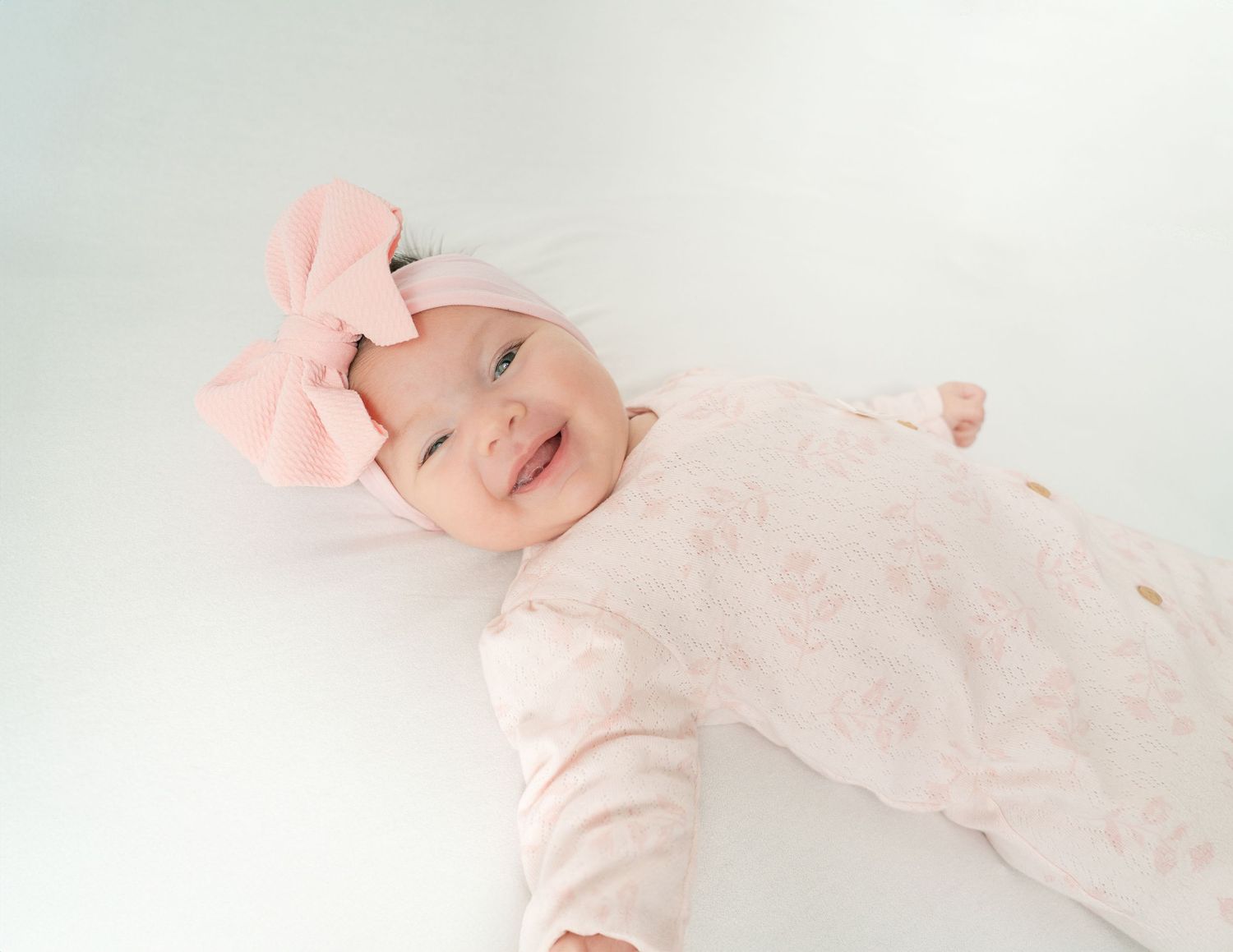 Newborn baby wearing soft pink outfit lying on white blanket in gentle natural lighting.