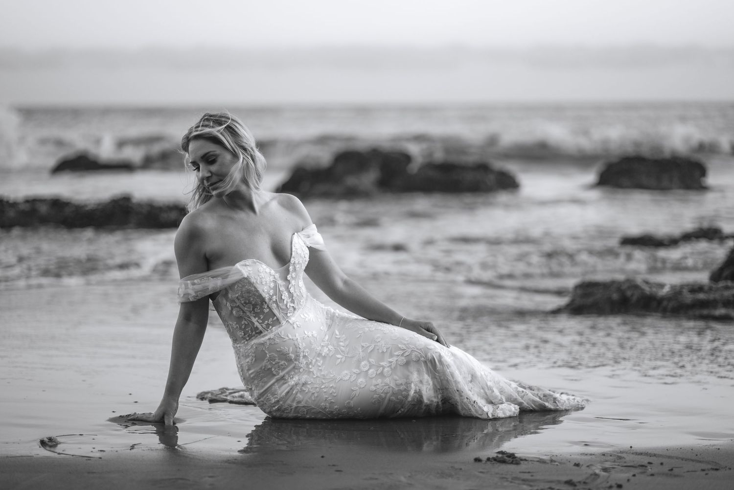 A person in a white dress lies gracefully on a sandy beach during a romantic photoshoot at sunset.
