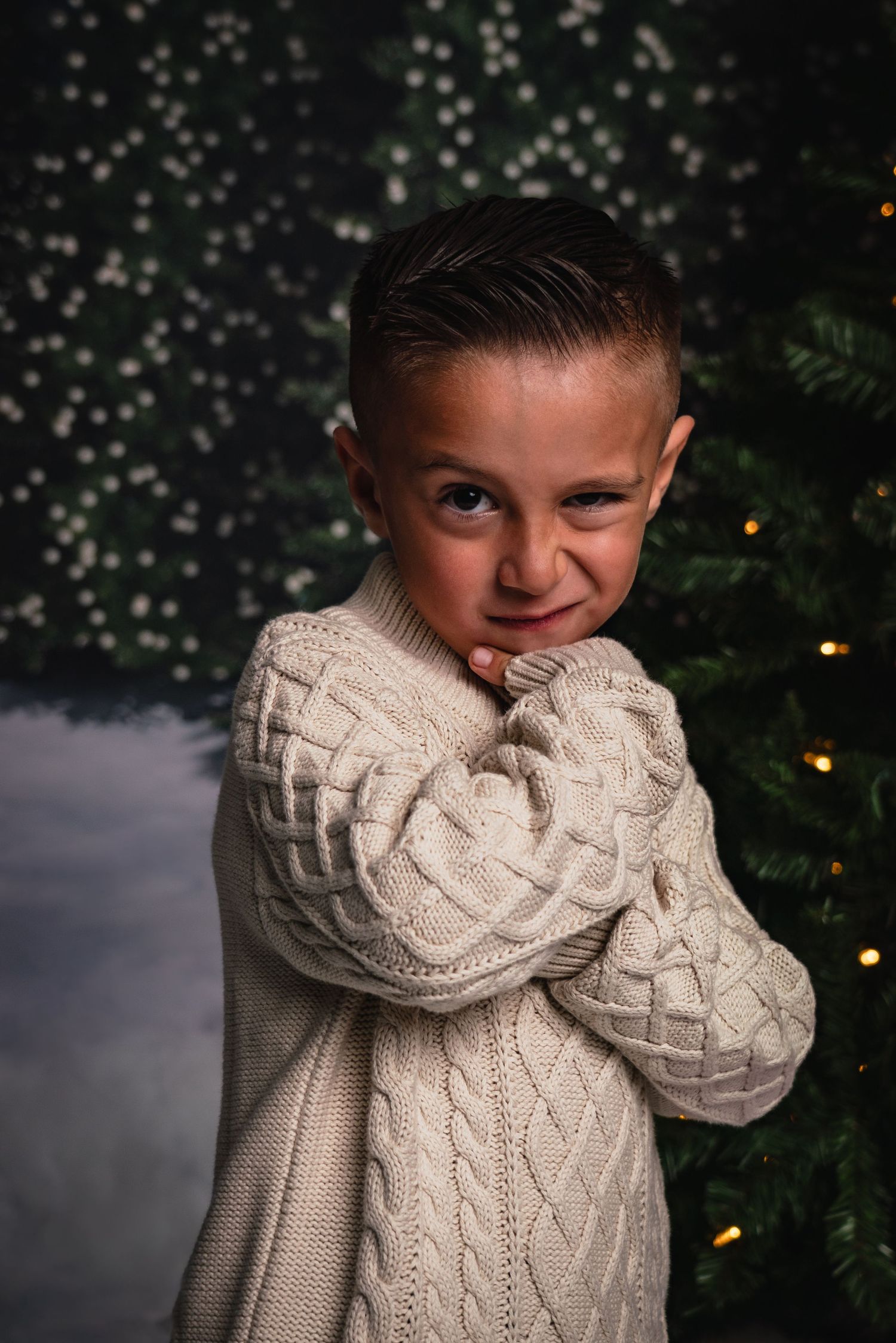 Child wearing a cozy white cable knit sweater poses playfully against a dark background with Christmas lights.