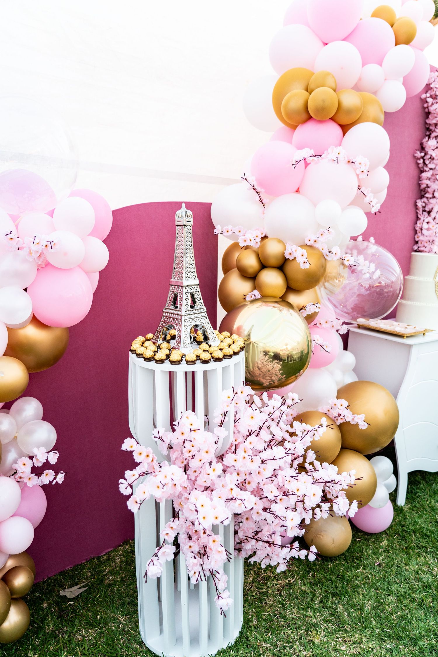 Pink and gold balloon arch with cherry blossoms and Eiffel Tower decorations on artificial grass.