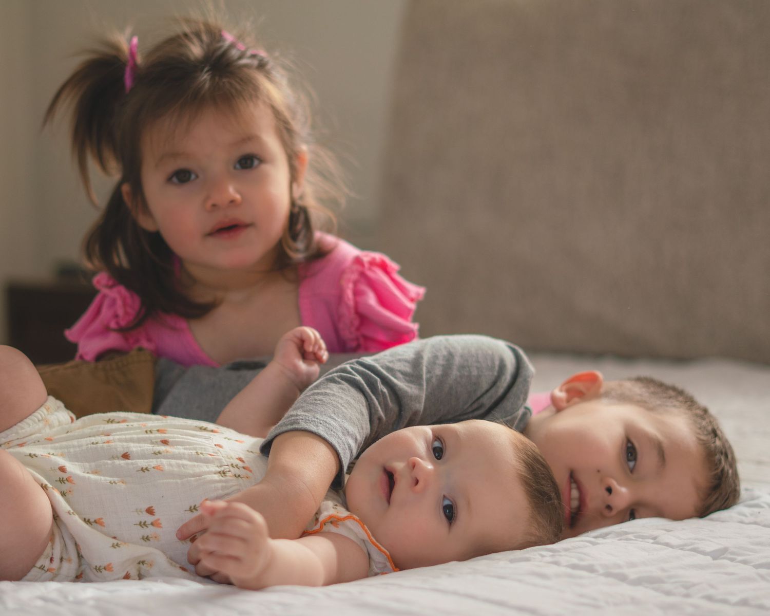 Three young babies and a toddler girl in pink dress lying together on a white bed in soft natural light.