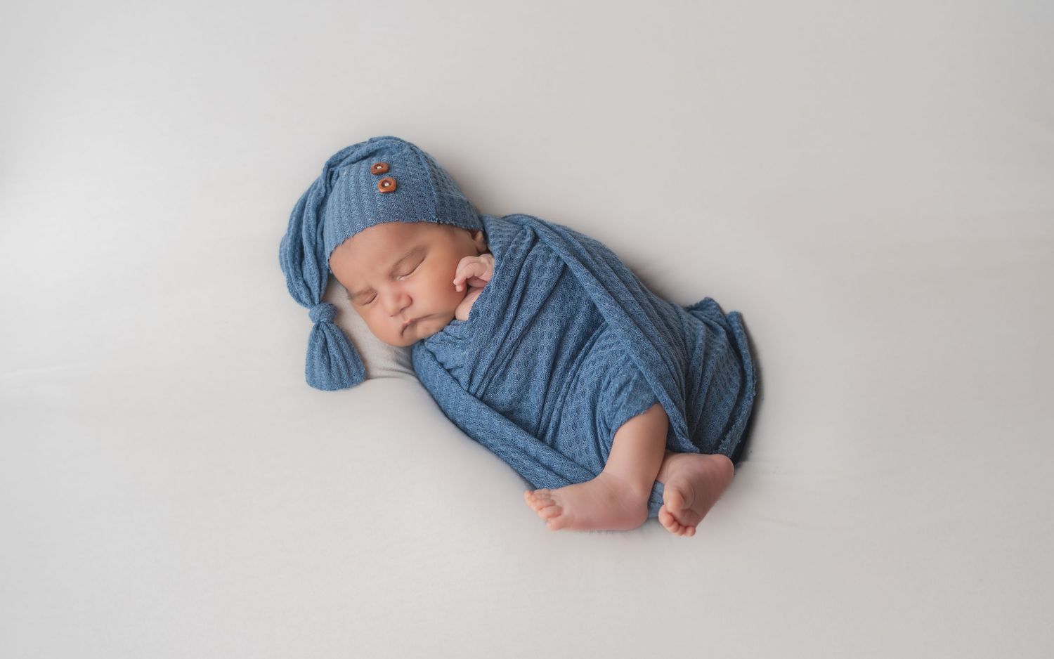 Newborn baby sleeping peacefully wrapped in a blue knit swaddle and matching hat on a white background.