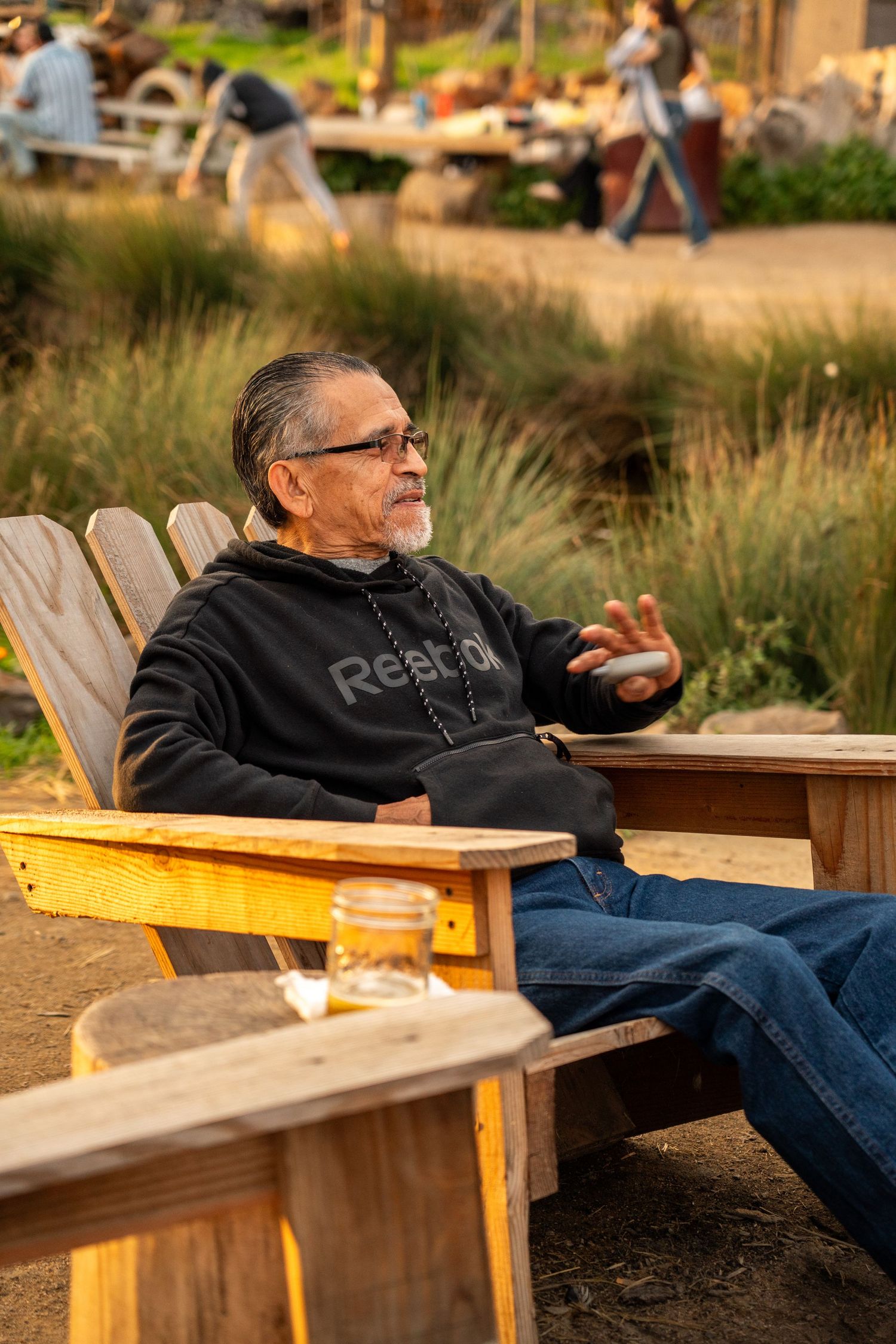 Older man in Reebok hoodie relaxing in wooden Adirondack chair outdoors at golden hour.